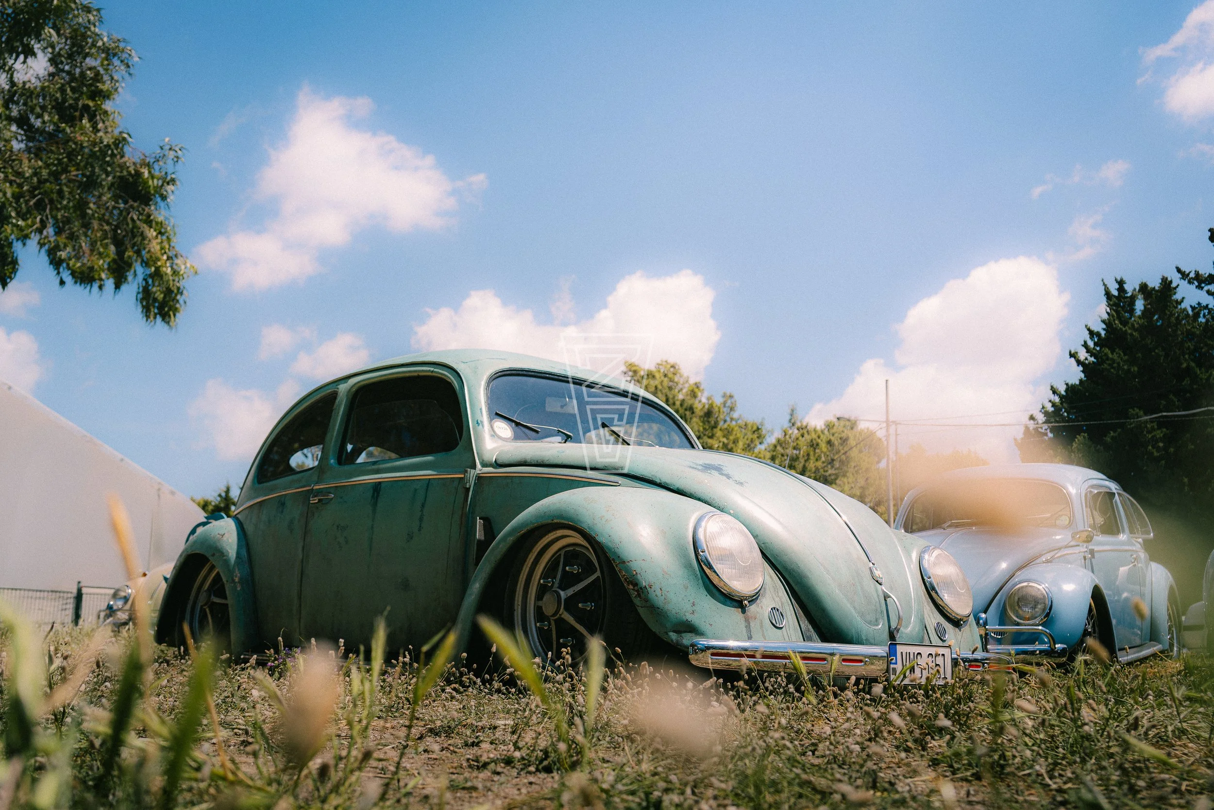 A vintage green car parked in a lot with other cars under a blue sky with some clouds.