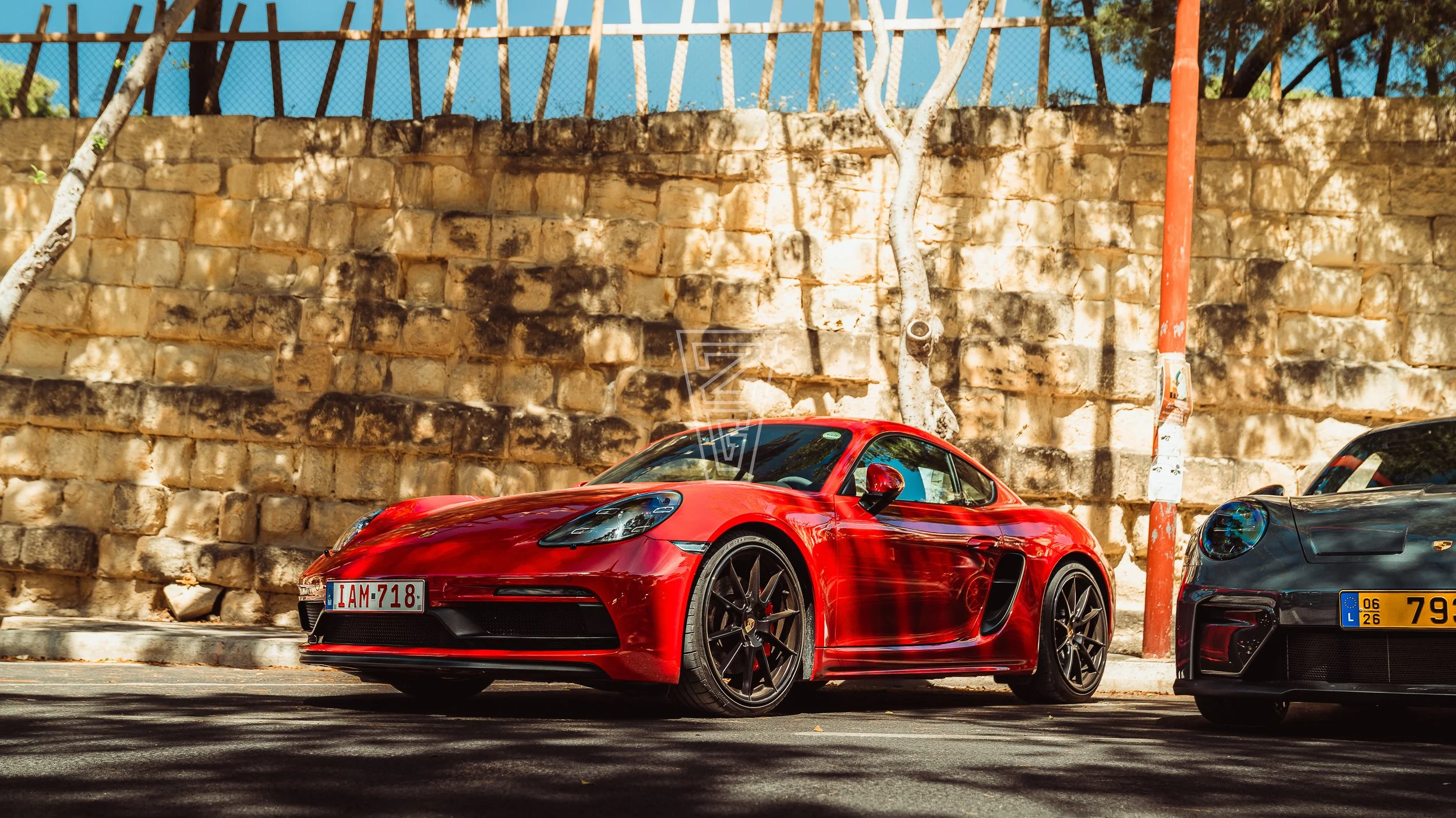 Red sports car parked beside a stone wall with trees overhead and a blue sky background.