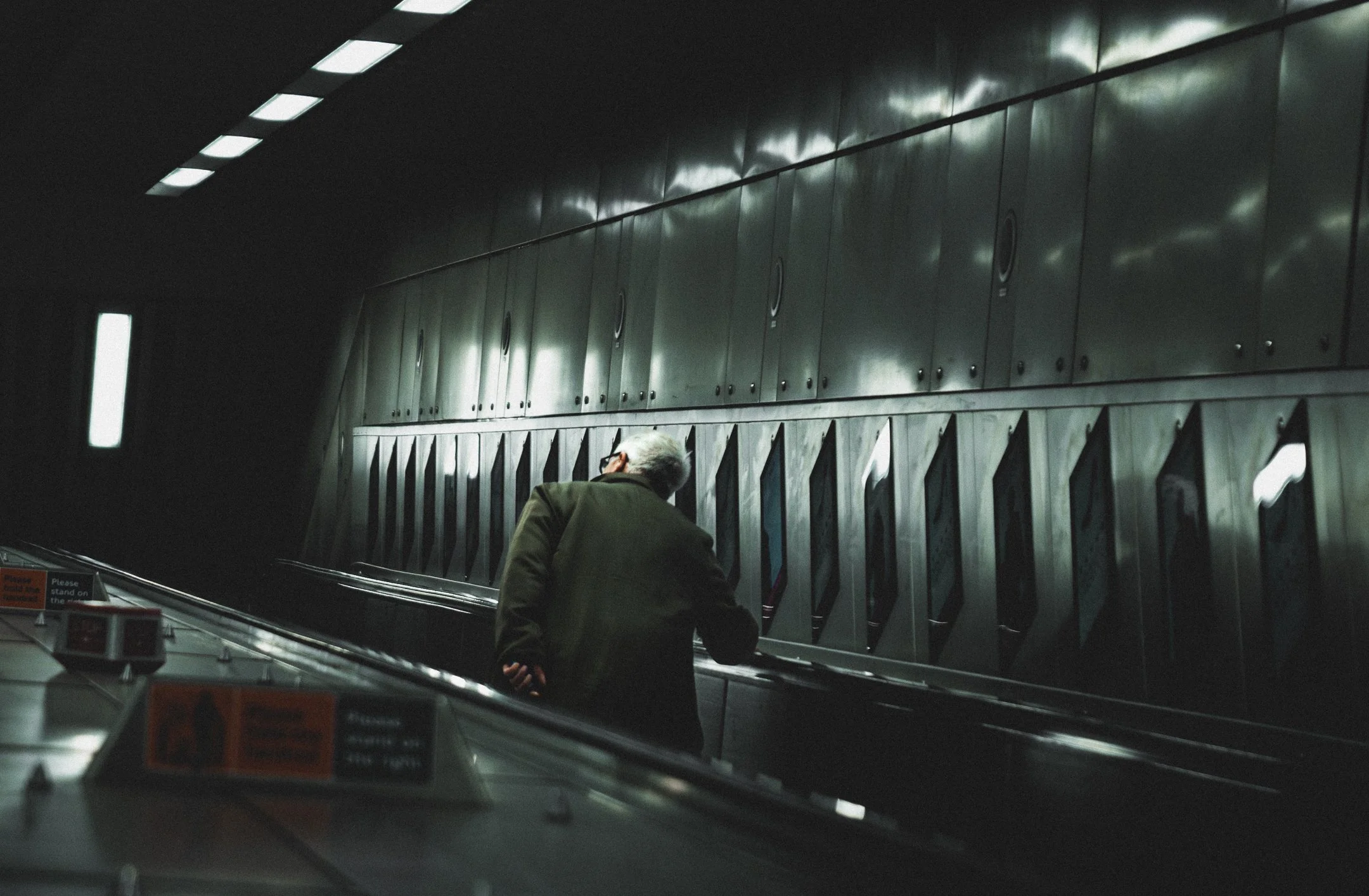 An elderly man on an escalator in a dimly lit subway station, with metallic walls and signage.