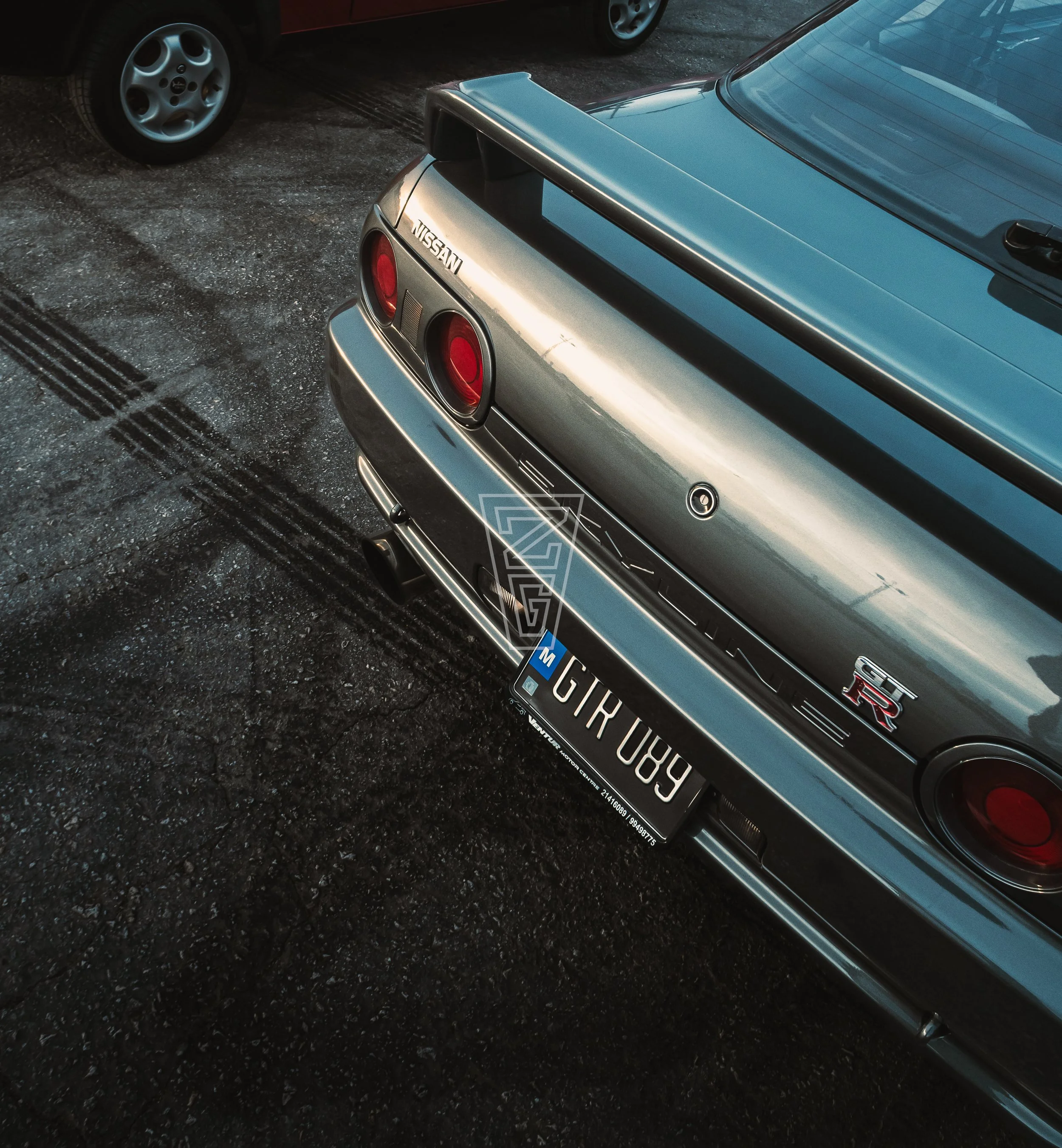 Close-up of the rear of a silver Nissan GTR sports car with a black spoiler, parked on a gravel surface, with part of another car visible in the background.