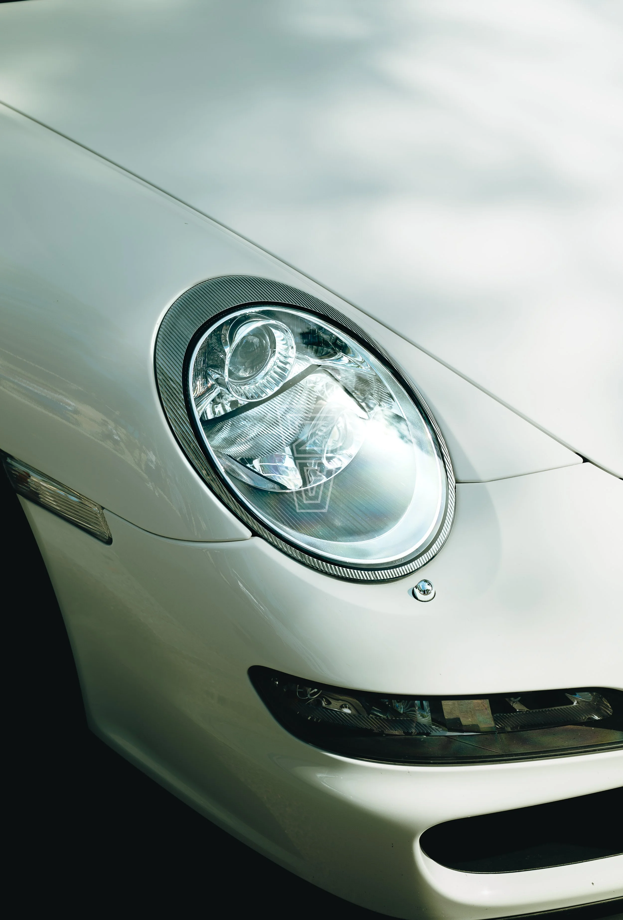 Close-up of a sleek white sports car's front, showing a round headlight, part of the hood, and a black grille.