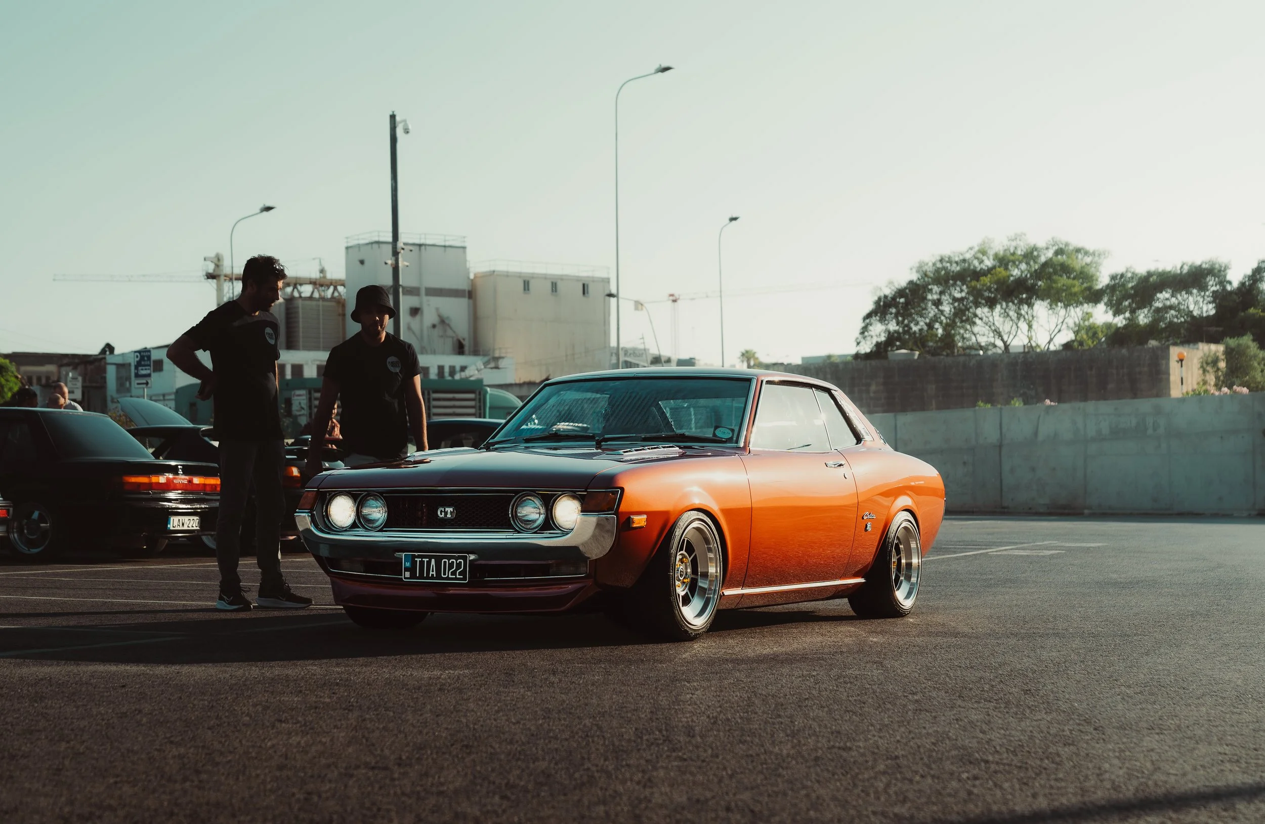Orange classic muscle car parked in a lot with two men standing nearby, one wearing a bucket hat, in front of industrial buildings and trees during daylight.