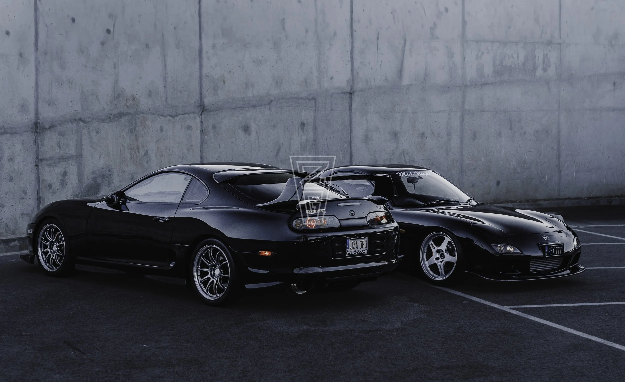 Two black sports cars parked side by side against a concrete wall, with custom rims and tinted windows.