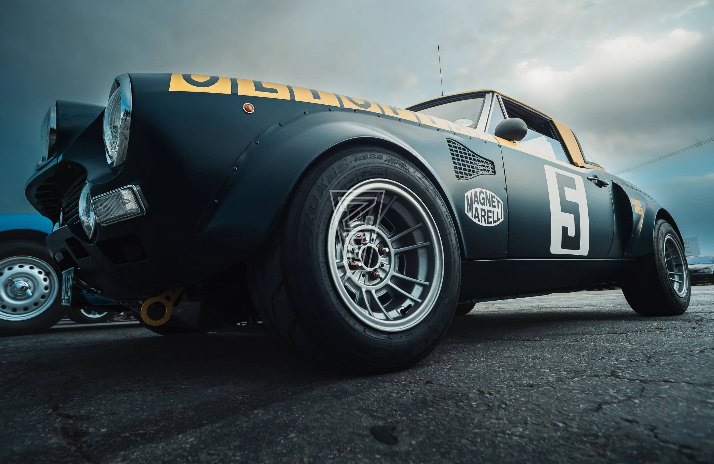Close-up of a vintage racing car with sponsor logos, including Magneti Marelli, showing large tires, a black and gold color scheme, and the number 5 on the side. Part of the front and side of the car is visible against a cloudy sky.