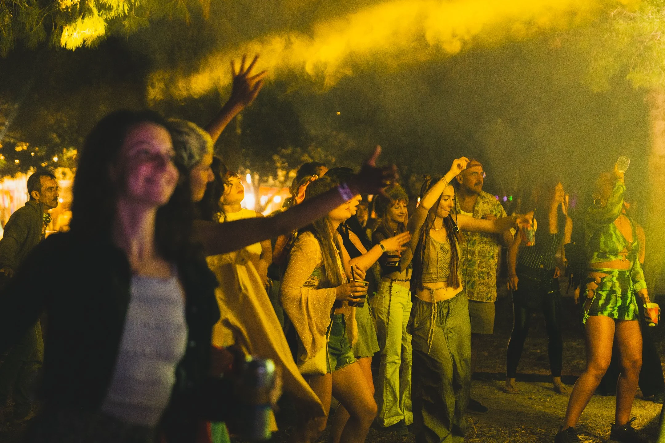 People dancing and enjoying music at an outdoor night event, illuminated by yellow lighting with trees in the background.