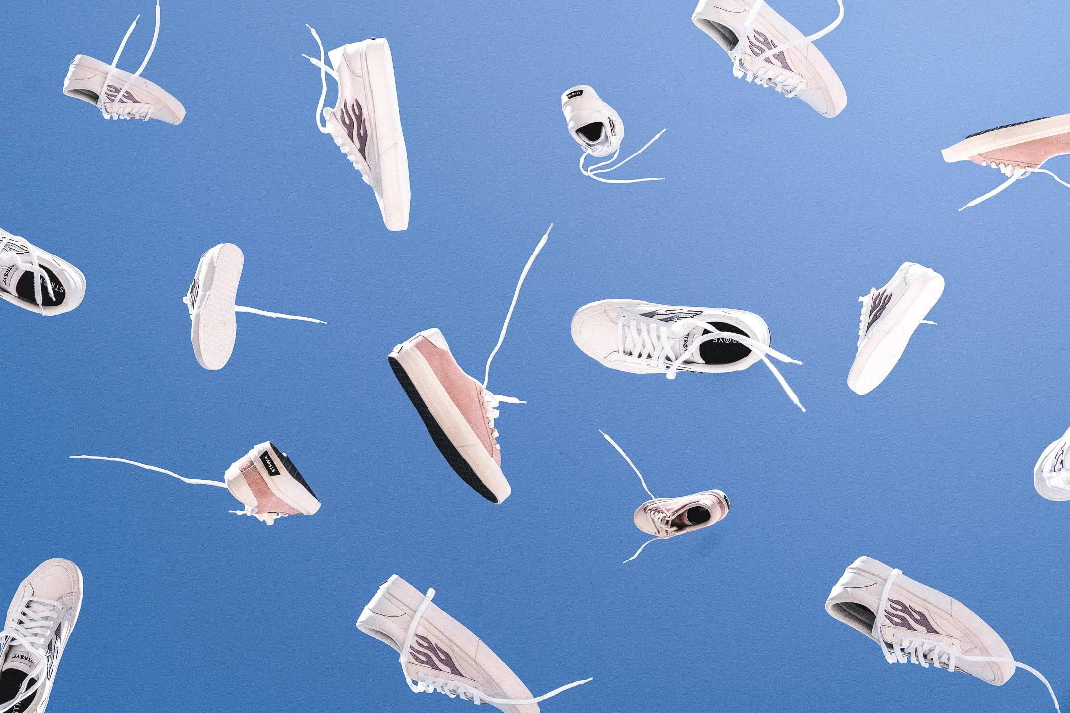 White and pink flame-patterned skate shoes suspended in the air against a clear blue sky.