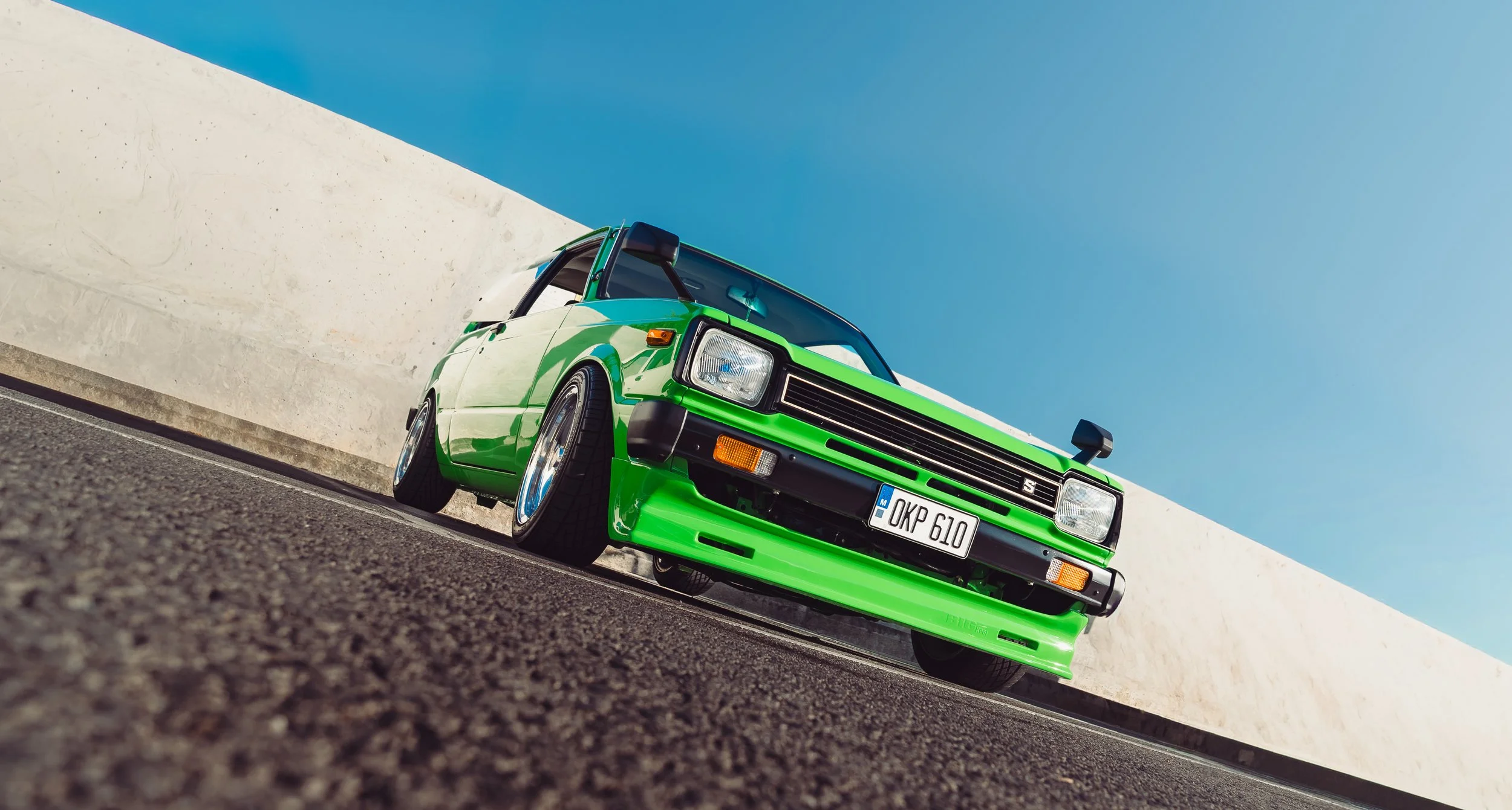 A bright green vintage car with a black roof parked on a paved surface next to a white concrete wall under a clear blue sky.
