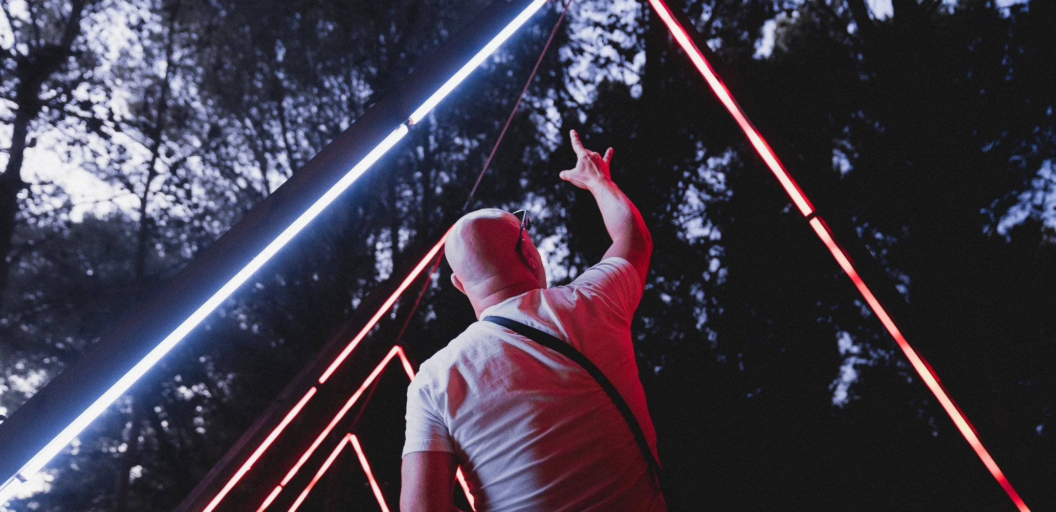 A bald man wearing glasses and a white t-shirt reaching up towards illuminated red and white neon light installation outdoors at dusk.