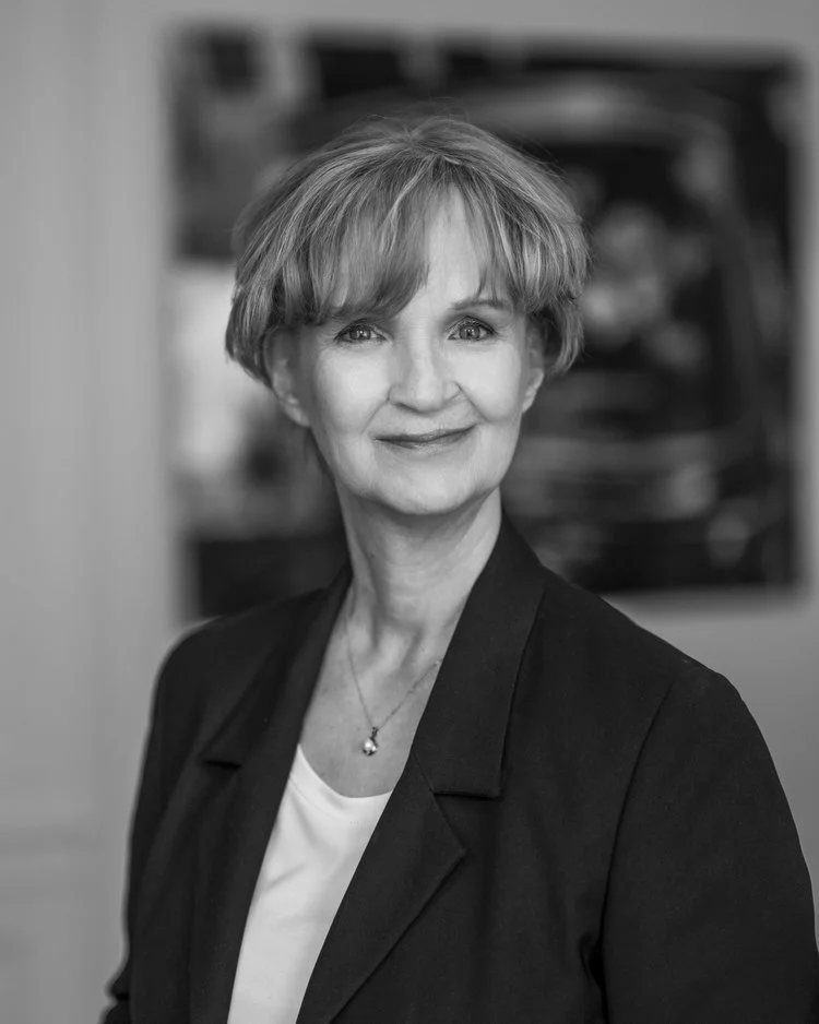 En svartvit porträttbild av en äldre kvinna med kort, lockigt hår, som bär en mörk blazer och ett vitt inre plagg, samt en liten halsdisk.