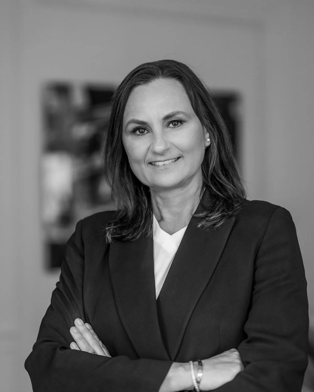 Svartvitt porträtt av en kvinna i affärskläder med korsade armar, leende, i ett kontorsmiljö.