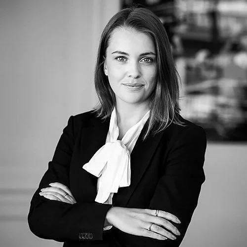 Kvinna i svart kostym och vit halsduk, porträtt i svartvitt, professionell miljö i bakgrunden.