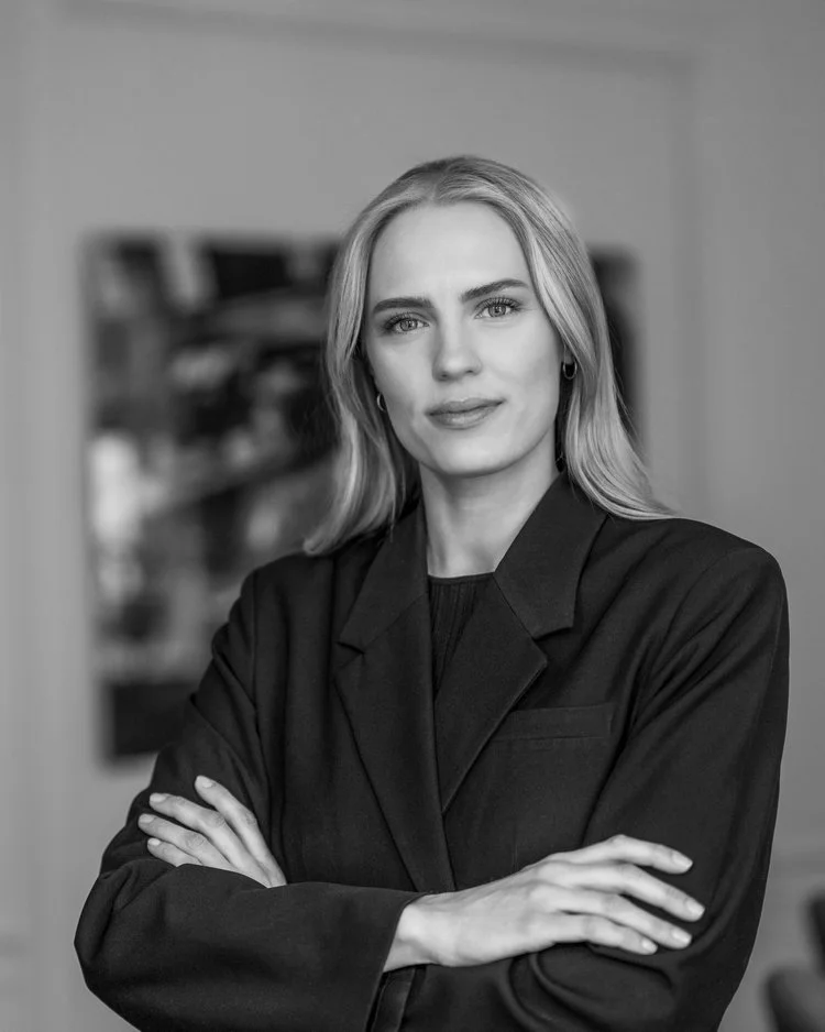 En ung kvinna med blont hår och ljusa ögon, klädd i en mörk kostym, står med korsade armar och tittar mot kameran, i ett svartvitt porträtt.