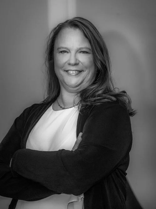 Kvinna med långt hår och ett leende, bär en svart jacka och ett vitt linne, står med armarna korsade.