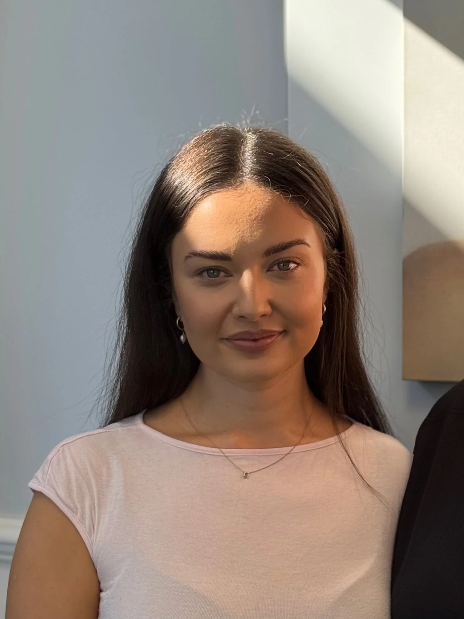 A young woman with long dark hair, light skin, and a subtle smile, standing in a room with light-colored walls and a wall-mounted lamp.