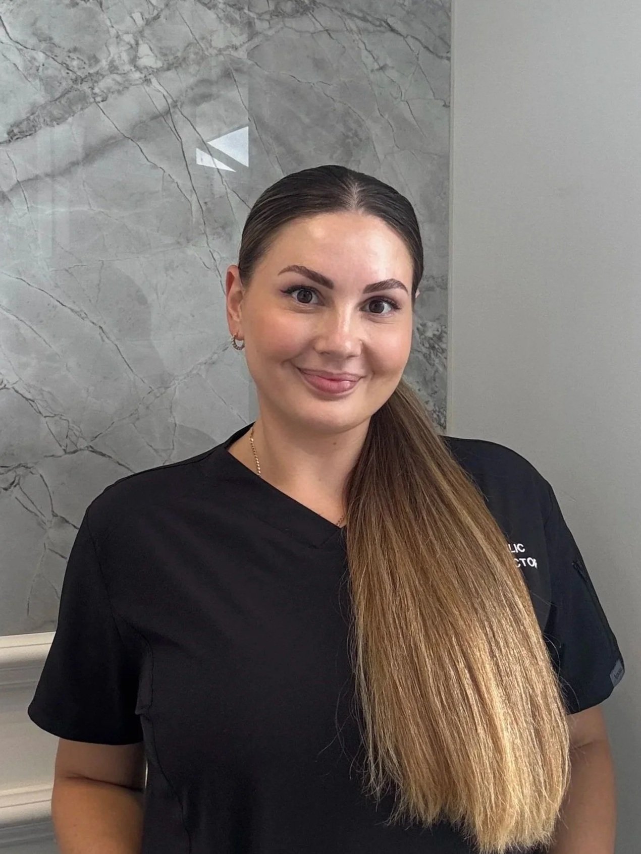 A woman with long, straight, light brown hair smiling, wearing a black medical uniform, standing indoors in front of a marble wall.