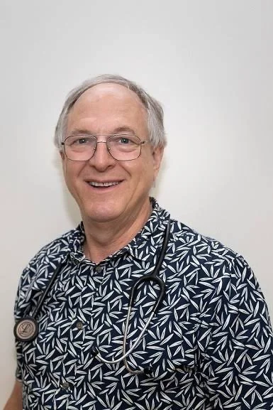 A smiling male doctor with glasses, wearing a patterned shirt and a stethoscope around his neck, standing against a plain background.