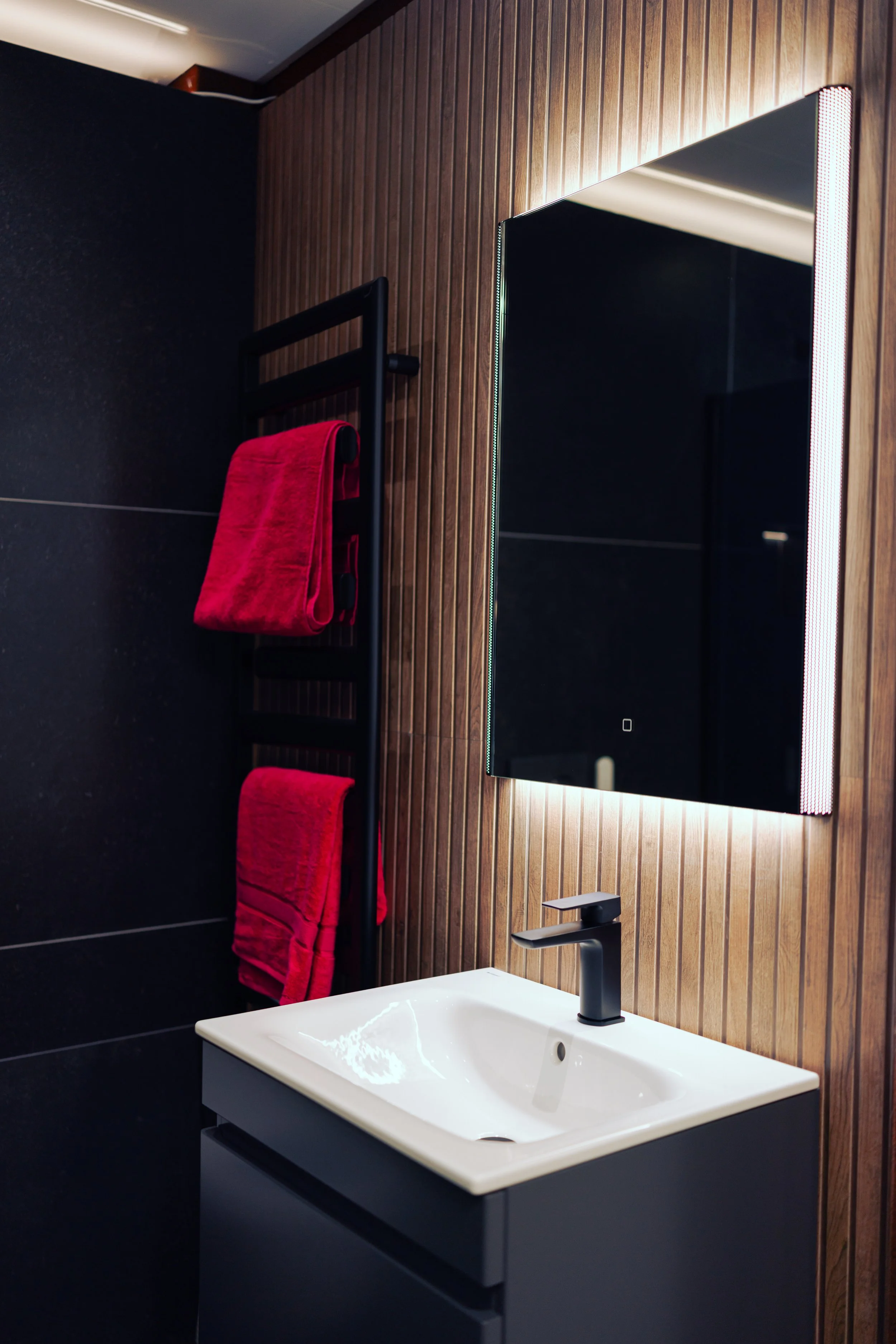 Modern bathroom with black tiled floor and wall, wooden paneling on one wall, a wall-mounted toilet, a black towel radiator, a small white sink with black vanity, and a mirror with LED lighting.