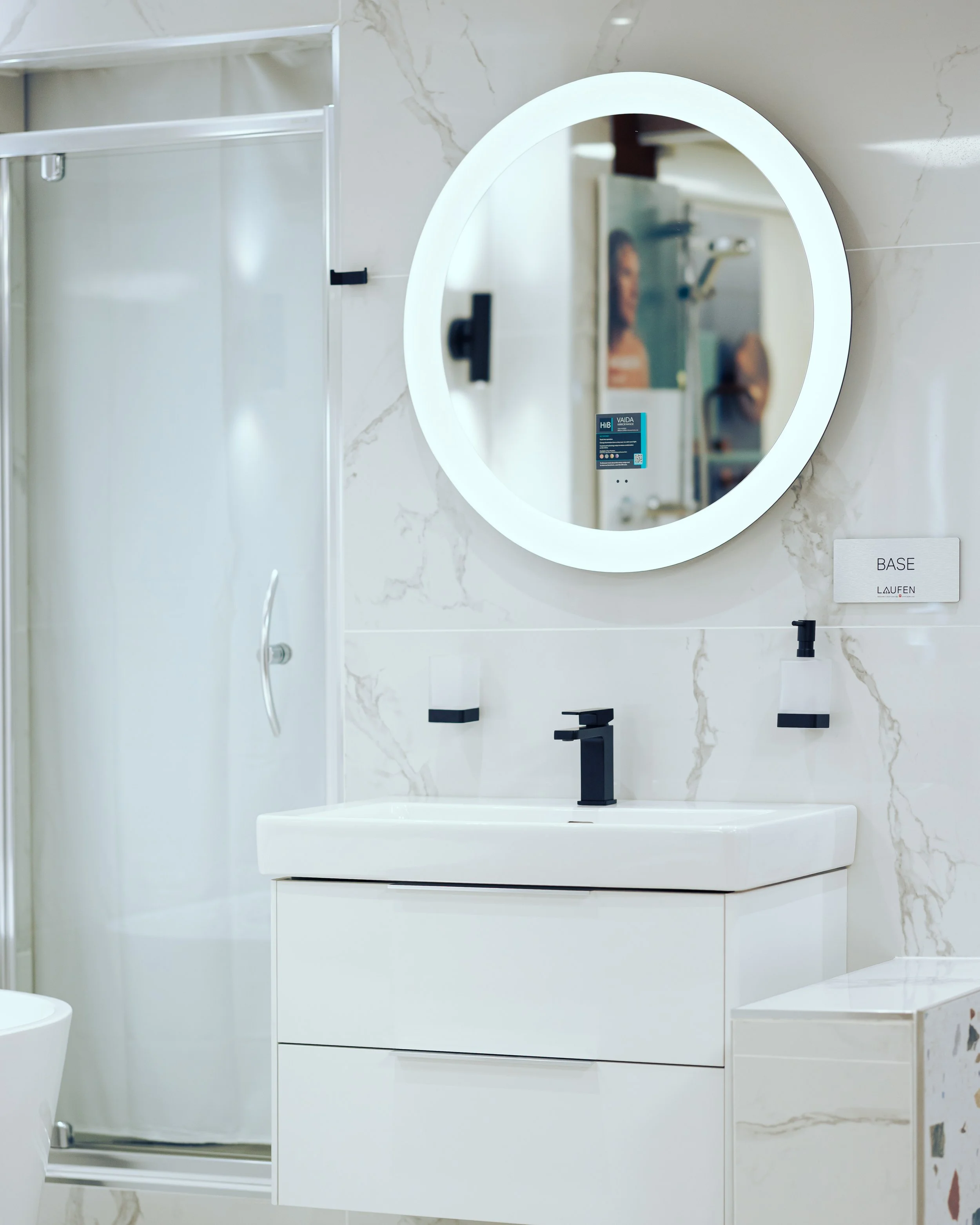 Modern bathroom with a white freestanding bathtub, wall-mounted toilet, marble tiled walls, a glass shower door, a white vanity with a round illuminated mirror, and black fixtures and accents.
