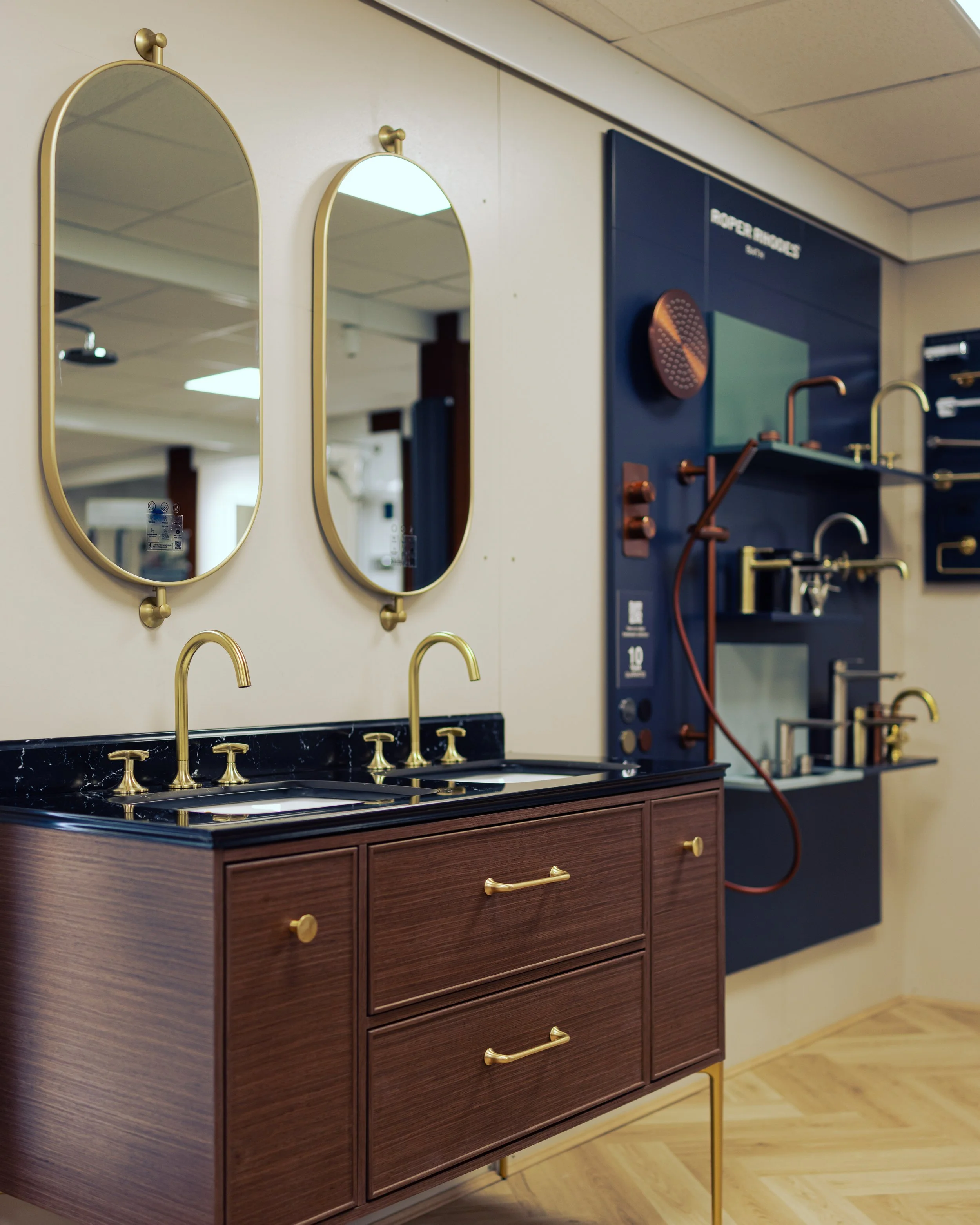 Bathroom vanity with a round white vessel sink and chrome faucet, pink rolled towels in open shelves, black brochures on a wooden countertop, and a mirror with LED lighting, all against dark gray marble wall tiles.