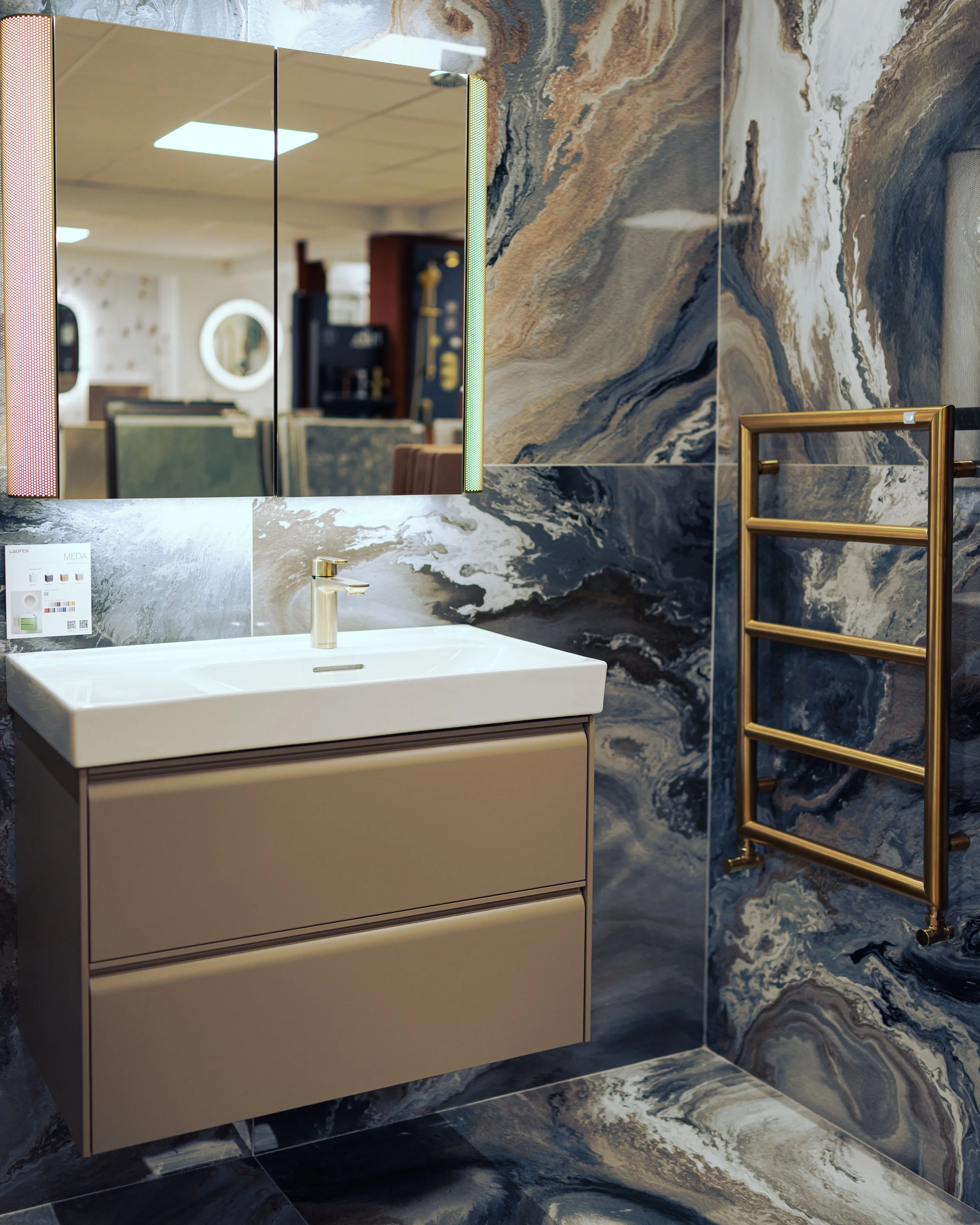 Restroom with a sign that reads 'Bayswater Bathrooms, City of Westminster,' a toilet with a blue seat cover, a dark blue bathroom vanity with a white countertop and sink, a mirror, and blue floral-patterned wallpaper.