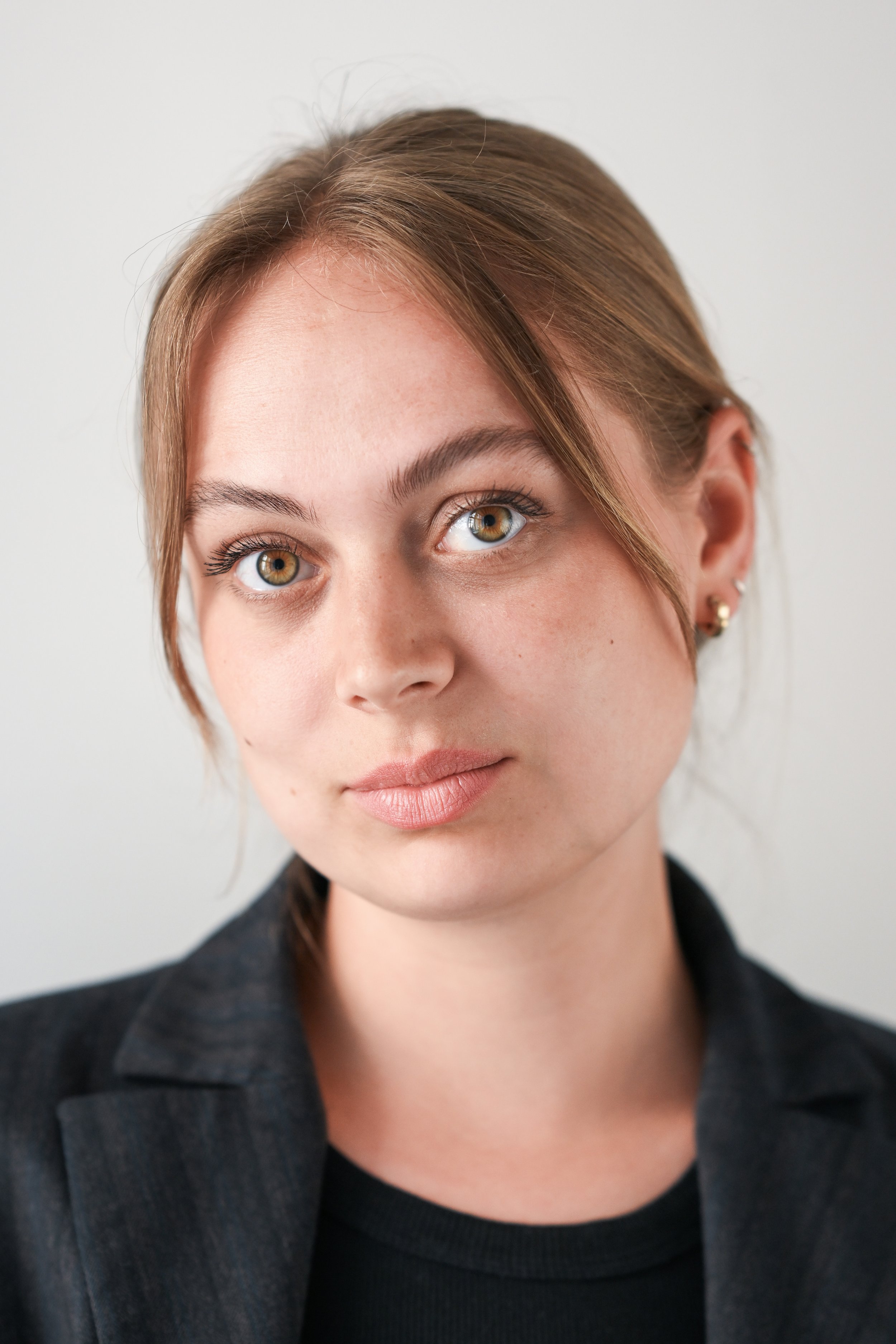 Portrætfoto af en ung kvinde med brunt mellem-til langt hår, grønne/gule øjne og let makeup, iført sort blazer og sort top, foran en neutral baggrund. Stemmecoach og co-founder af Voice Performance Collective.