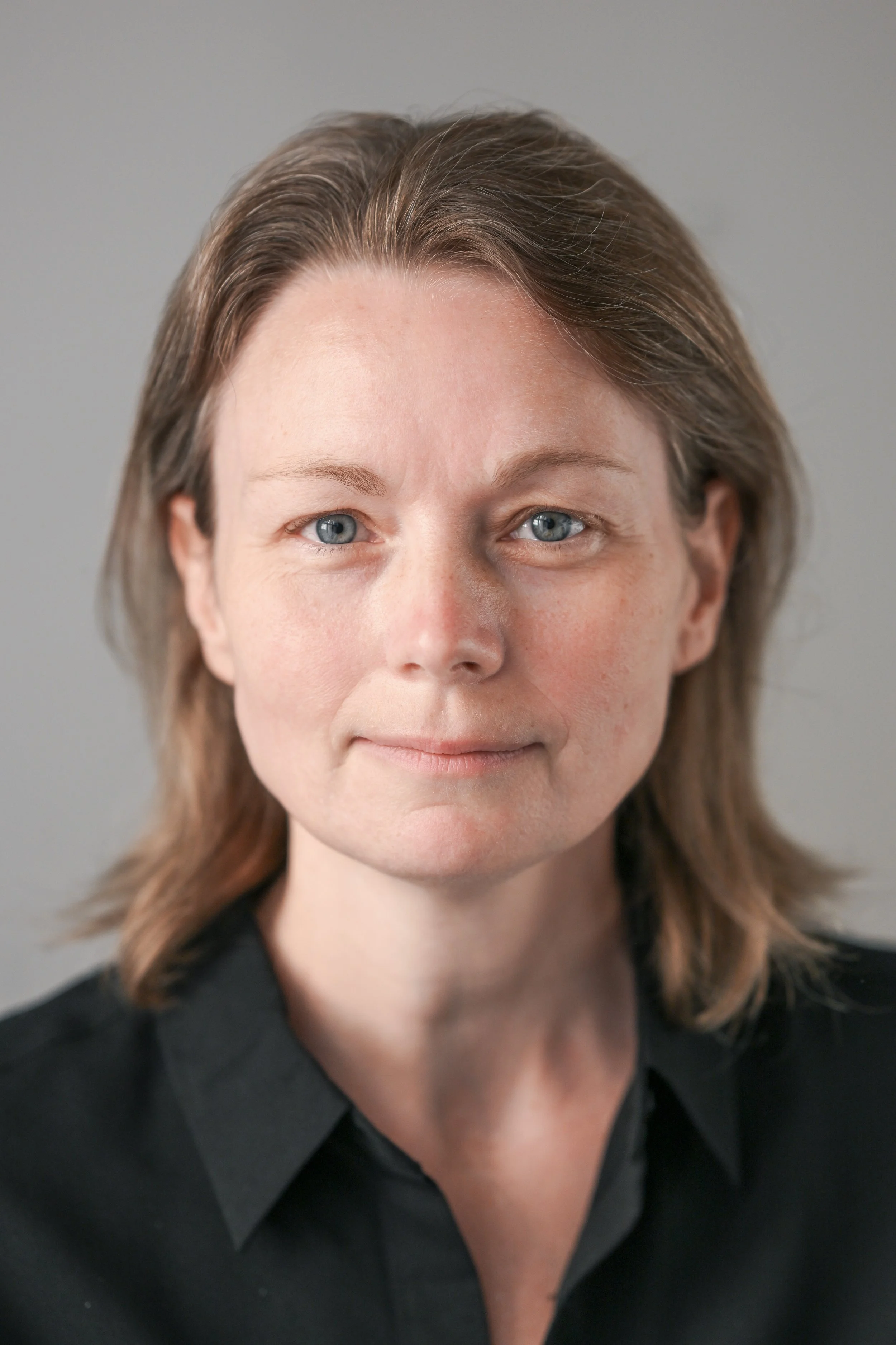 Portræt af en kvinde med brunt, skulderlangt hår, blå øjne og en sort skjorte, foran en grå baggrund. Stemmecoach og co-founder af Voice Performance Collective.