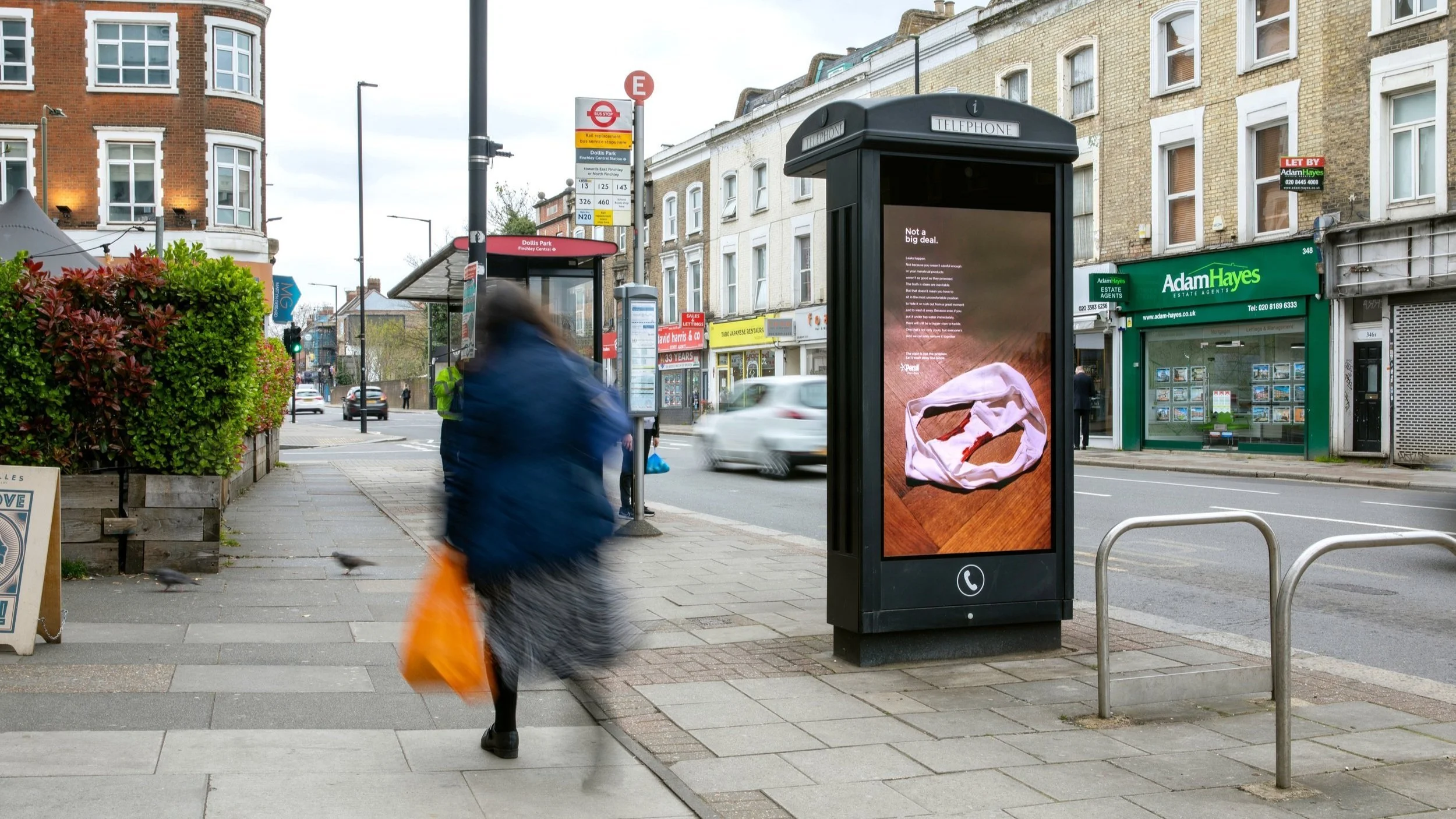 People walking past a bus stop and digital advertising kiosk on a city street, with shops and buildings in the background.