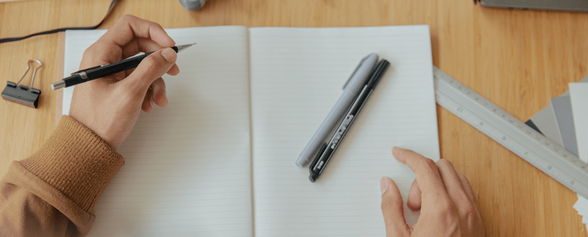 une personne tient un stylo à bille au-dessus d'un cahier ouvert à lignes, avec deux autres marqueurs sur la page et des outils de dessin à proximité sur une surface en bois.