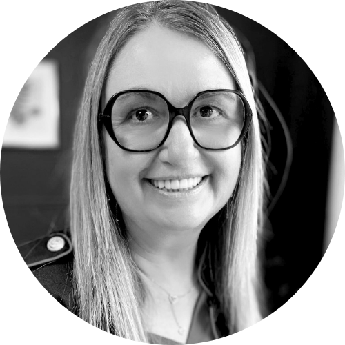 Une femme souriante portant de grandes lunettes rondes et une chemise noire, avec des cheveux longs et clairs, en noir et blanc.