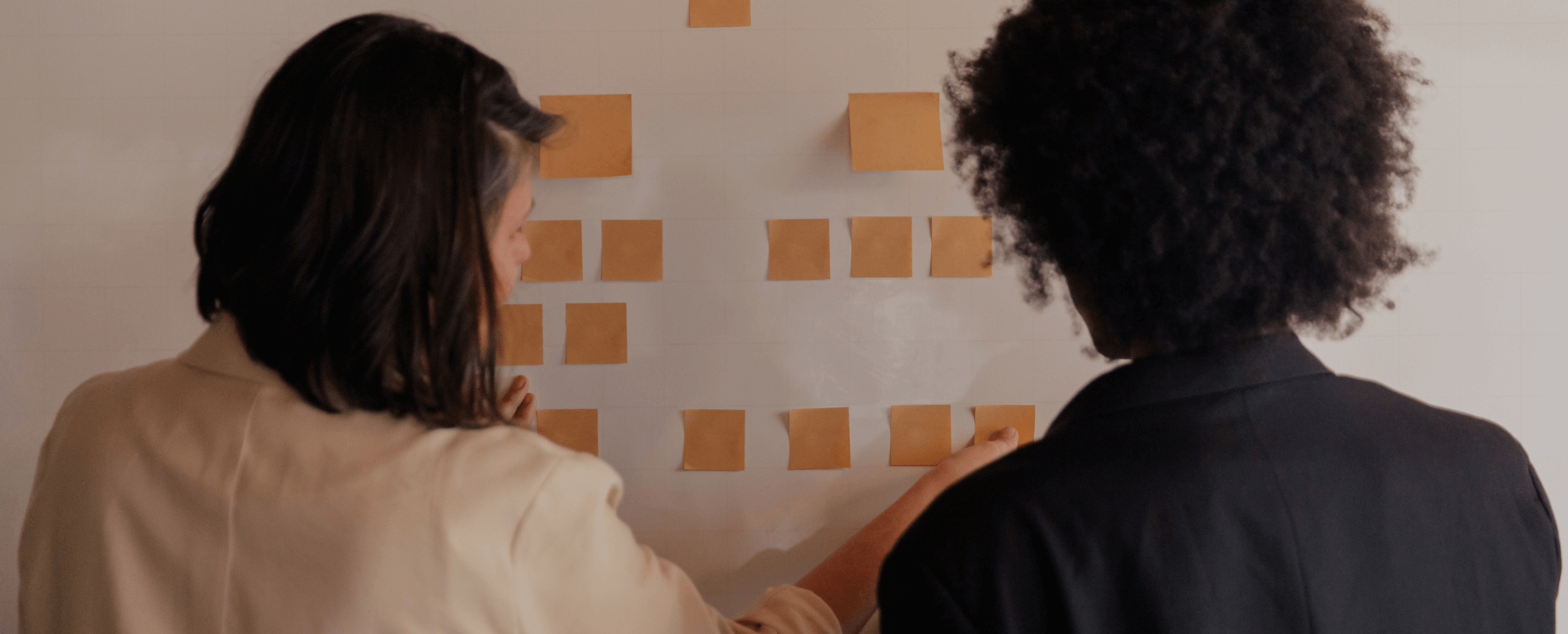 Deux femmes regardent un tableau avec des notes adhésives en jaune sur un mur blanc.