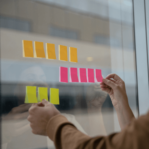 Une personne colle des notes adhésives de couleurs différentes (orange, rose, jaune) sur une vitre.