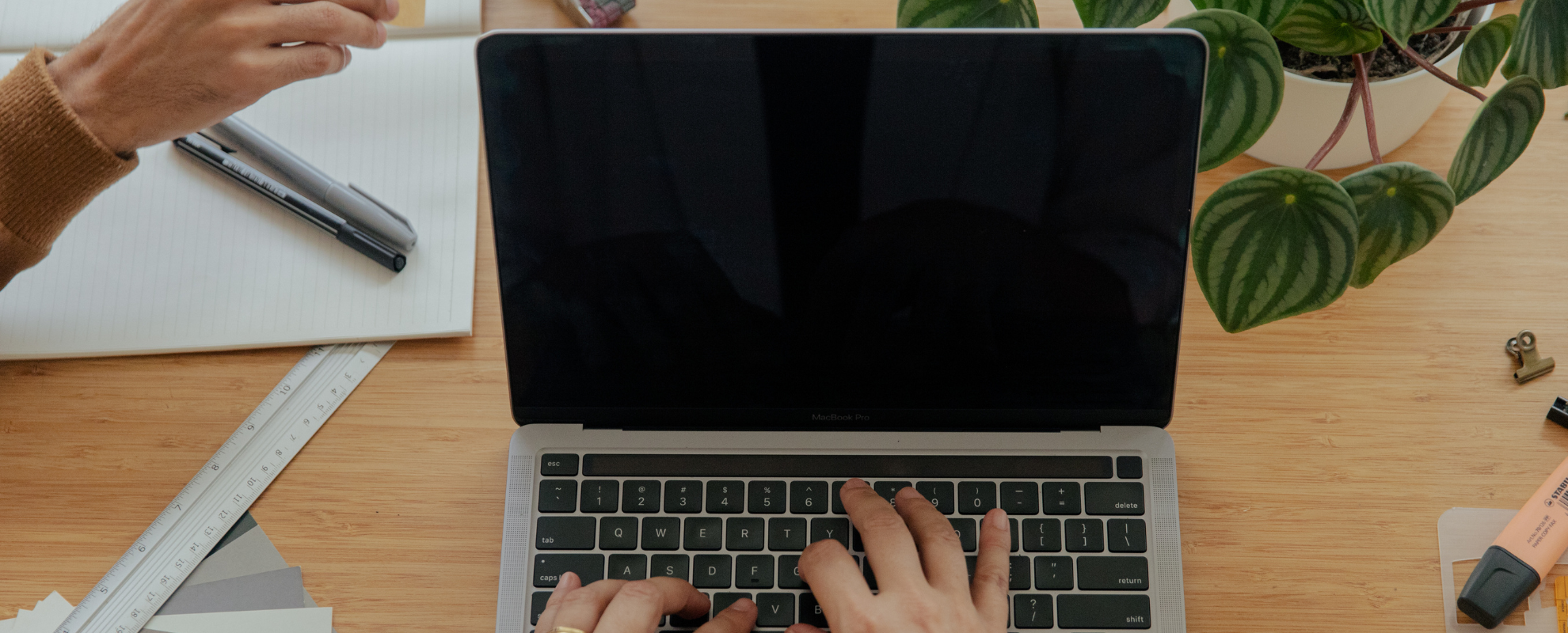 Une personne utilisant un ordinateur portable sur un bureau en bois, avec une plante à côté, un cahier, un stylo, une règle, une pince et un tube de colle.