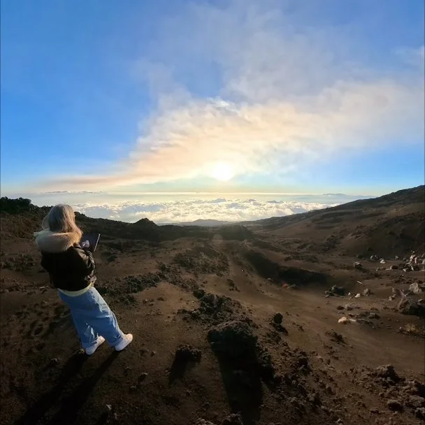 Artist Medea standing on the top of Etna Volcano, looking at the sunset above the clouds, holding tablet and life sketching on it the rugged landscape.