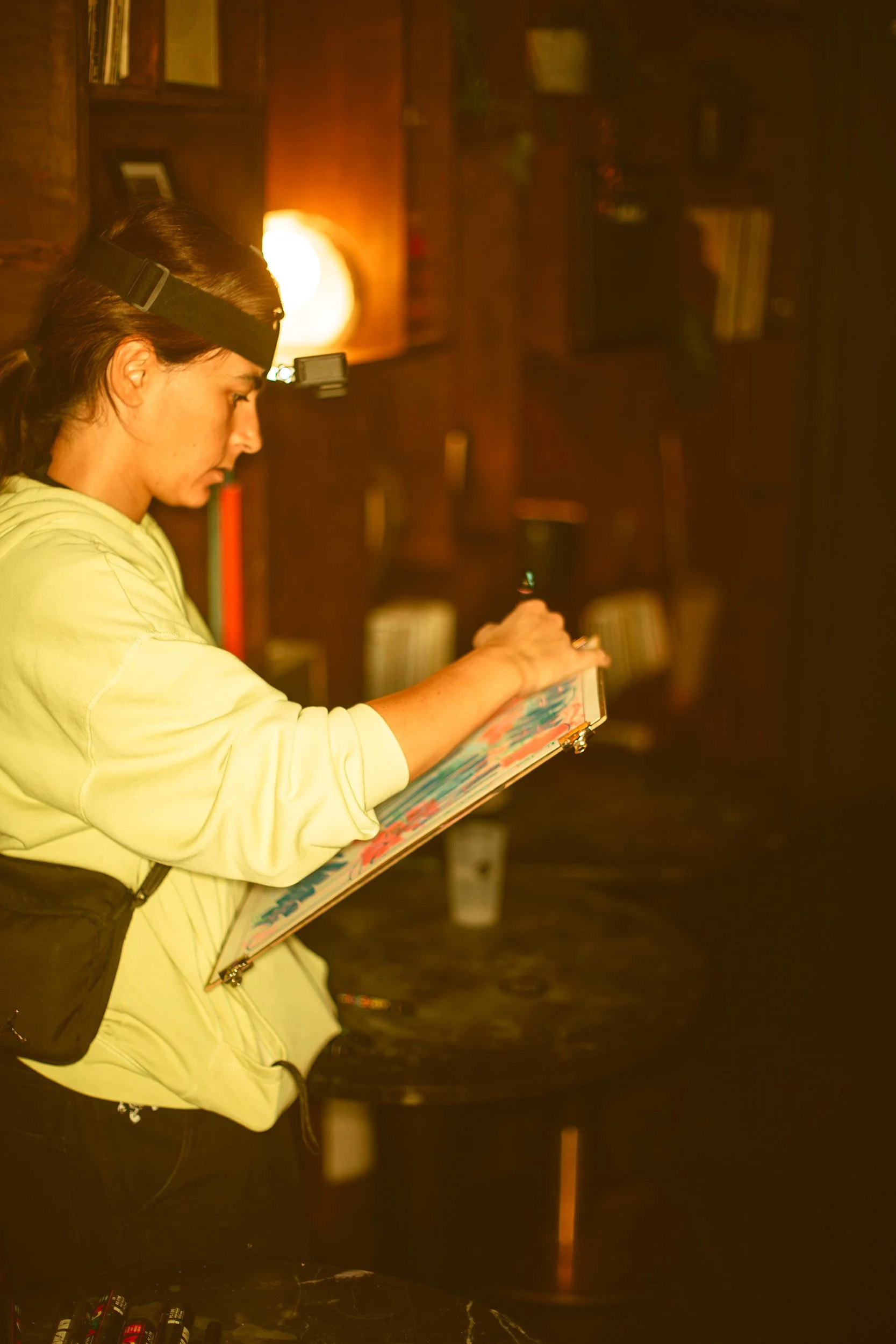 A young woman with dark hair which is Artist Medea wearing a GoPro Camera on her forehead and a yellow hoodie, holding a board with paper on it on which she is live sketching in a jazz club with wooden shelves and books in the background.
