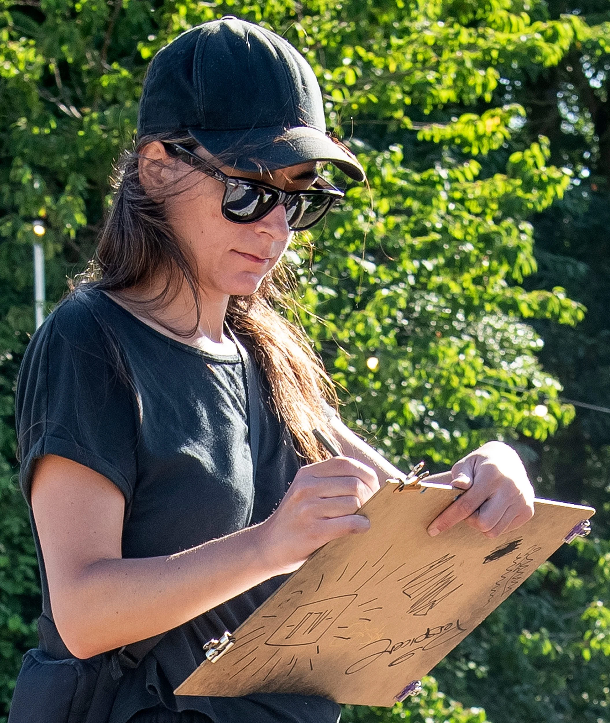 A woman which is Artist Medea wearing a black cap and sunglasses is outdoors, drawing with black marker on a paper supported by MDF Board.