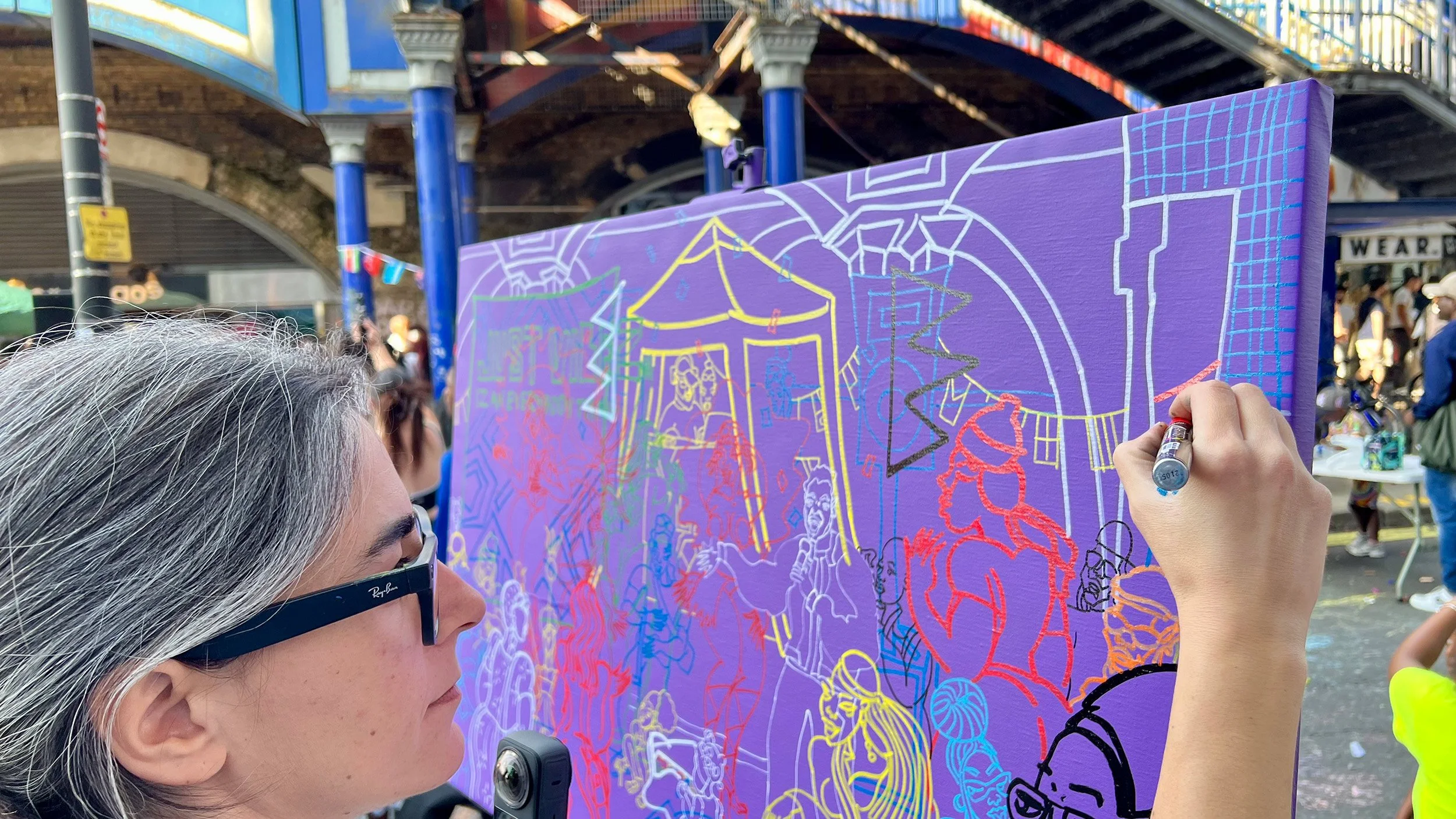 Person with gray hair and sunglasses painting colorful graffiti on a purple canvas at an outdoor event with a crowd and bridge in the background.