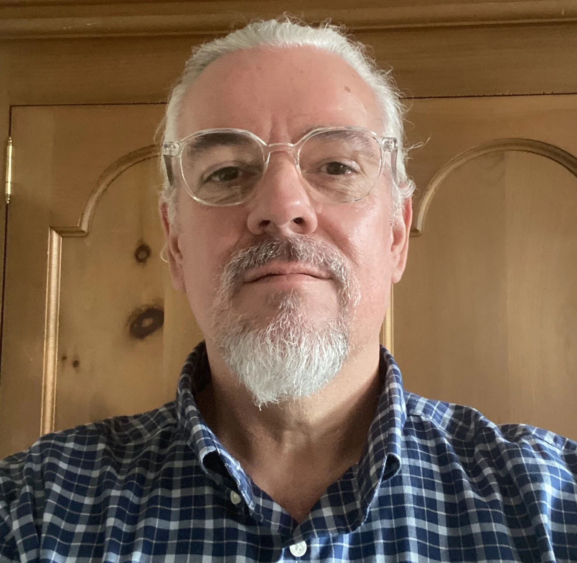 A middle-aged man with white hair, a white beard and mustache, wearing clear glasses and a navy blue checkered shirt, standing in front of a wooden door.