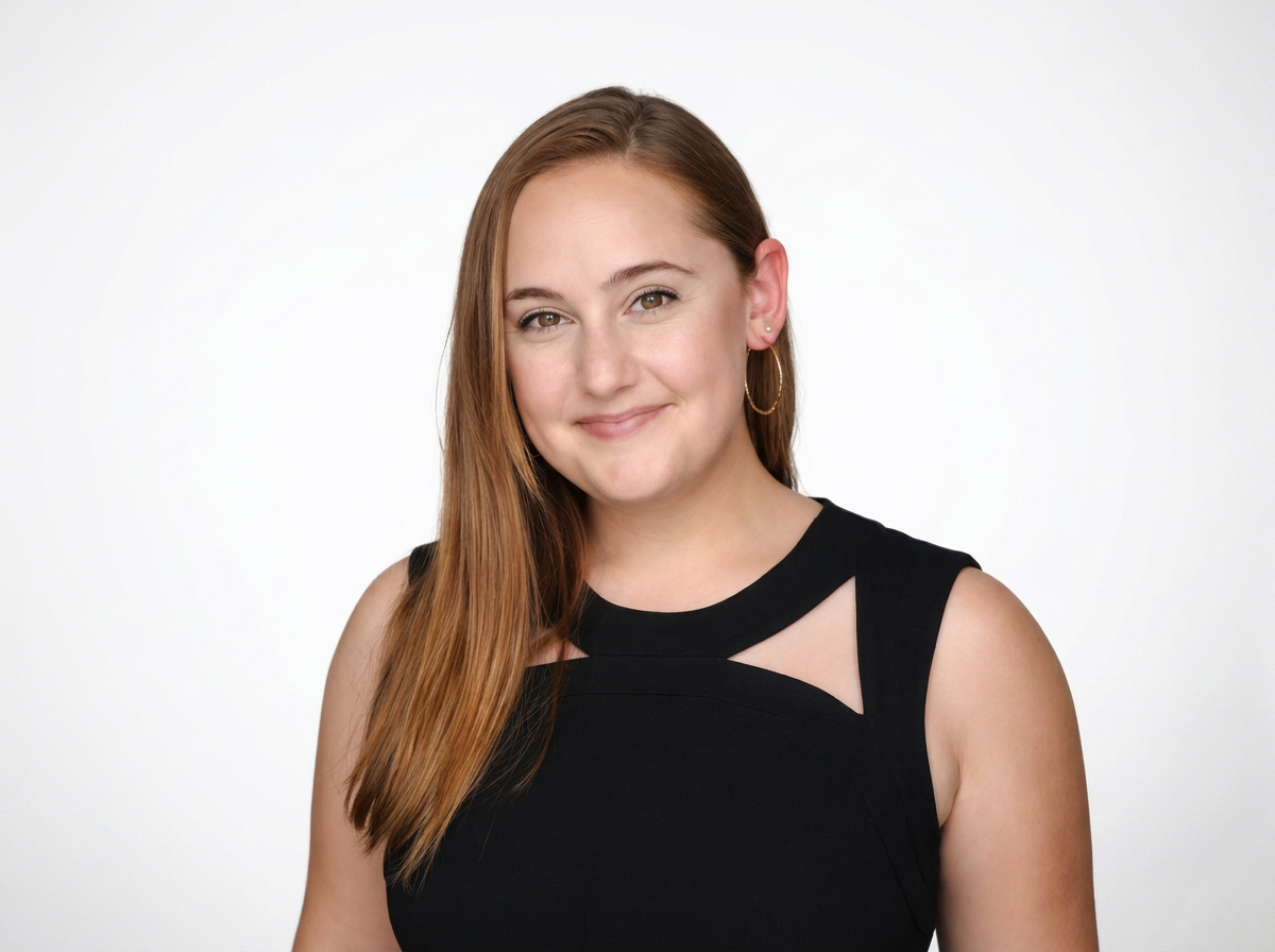 Headshot of a young woman with long red hair, smiling, wearing a black sleeveless top with cut-out details, and gold hoop earrings, against a plain white background.