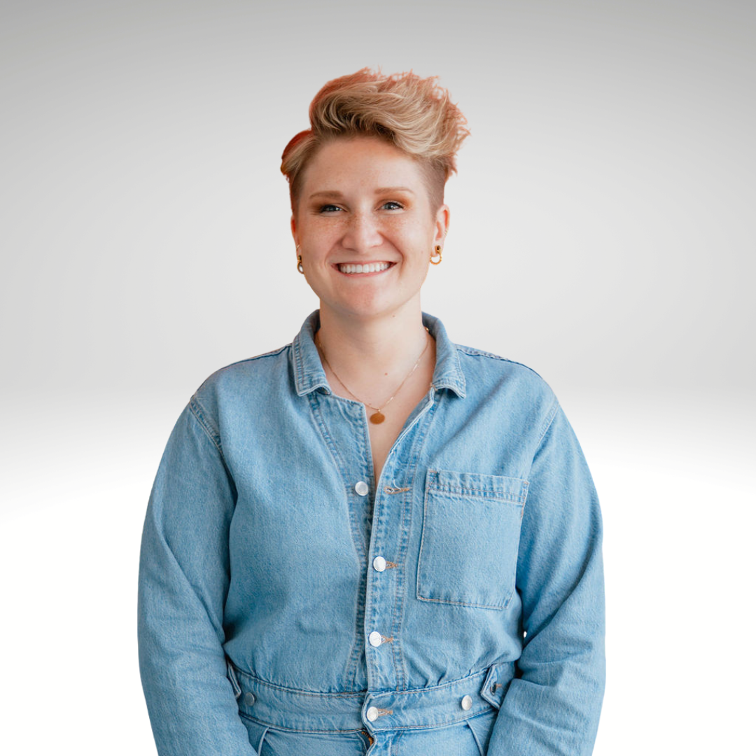 A woman with short, tousled blonde hair smiling and wearing a denim jacket and gold jewelry, standing against a plain light background.