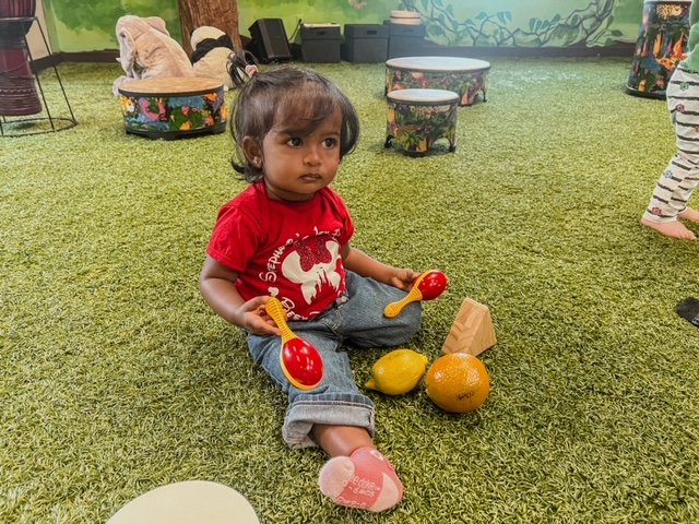 A young girl sitting on artificial grass holding red and yellow toy maracas, with orange and yellow fruits and a small wooden block in front of her, in an indoor play area with cushions and other children in the background.