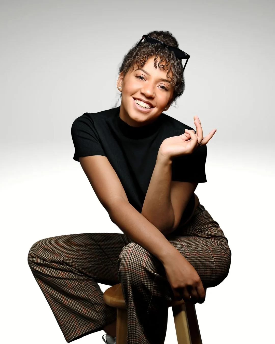 A vocal coach and piano teacher with curly hair, wearing a black t-shirt and plaid pants, sitting on a wooden stool, smiling, with sunglasses on her head and making a casual hand gesture.