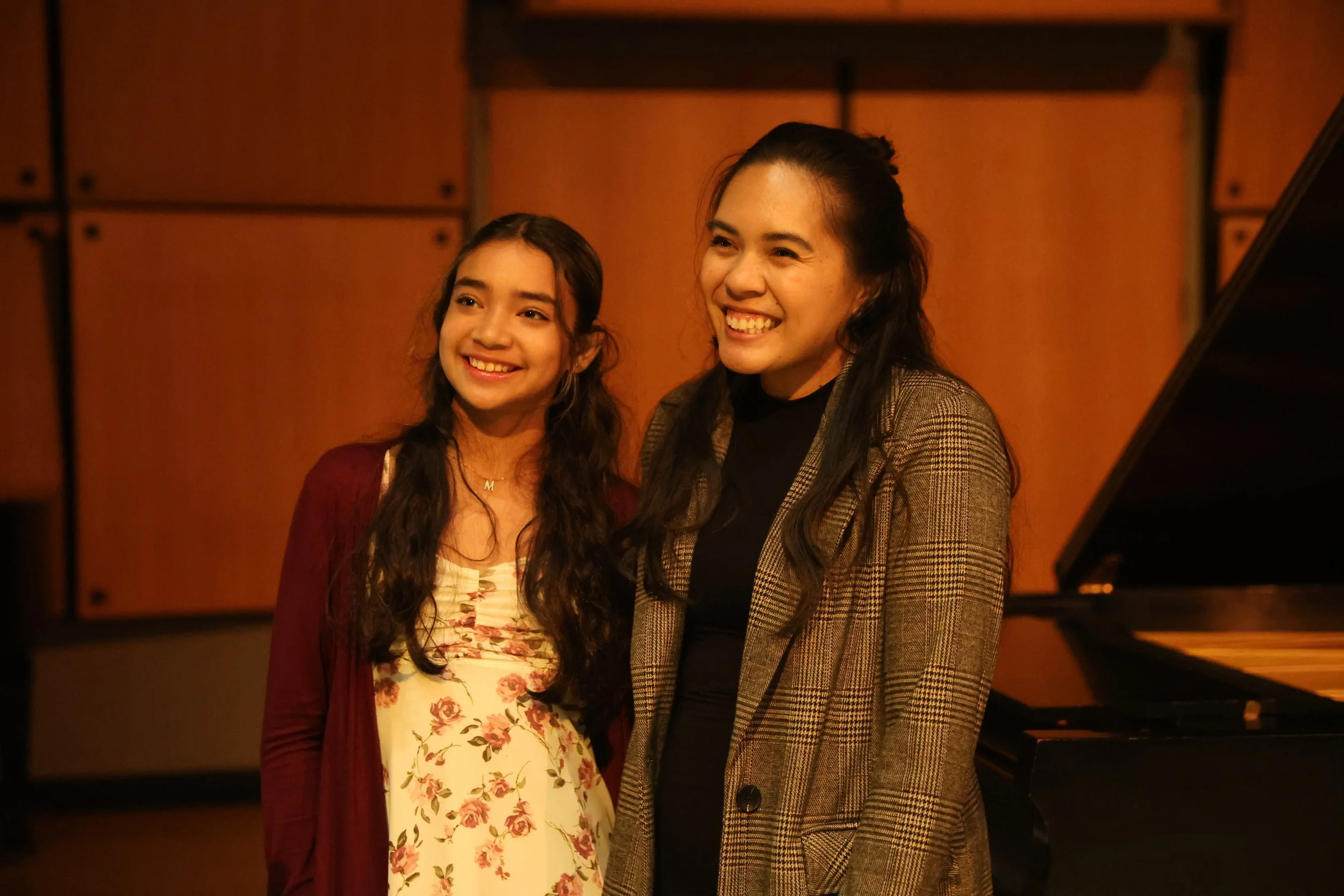 Two women standing together smiling in a warmly lit room with a piano visible on the right.