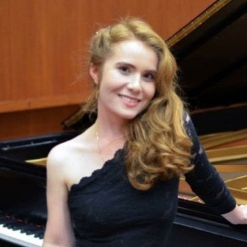 A young woman with long, curly red hair, wearing a black one-shoulder dress, sitting at a grand piano and smiling at the camera.
