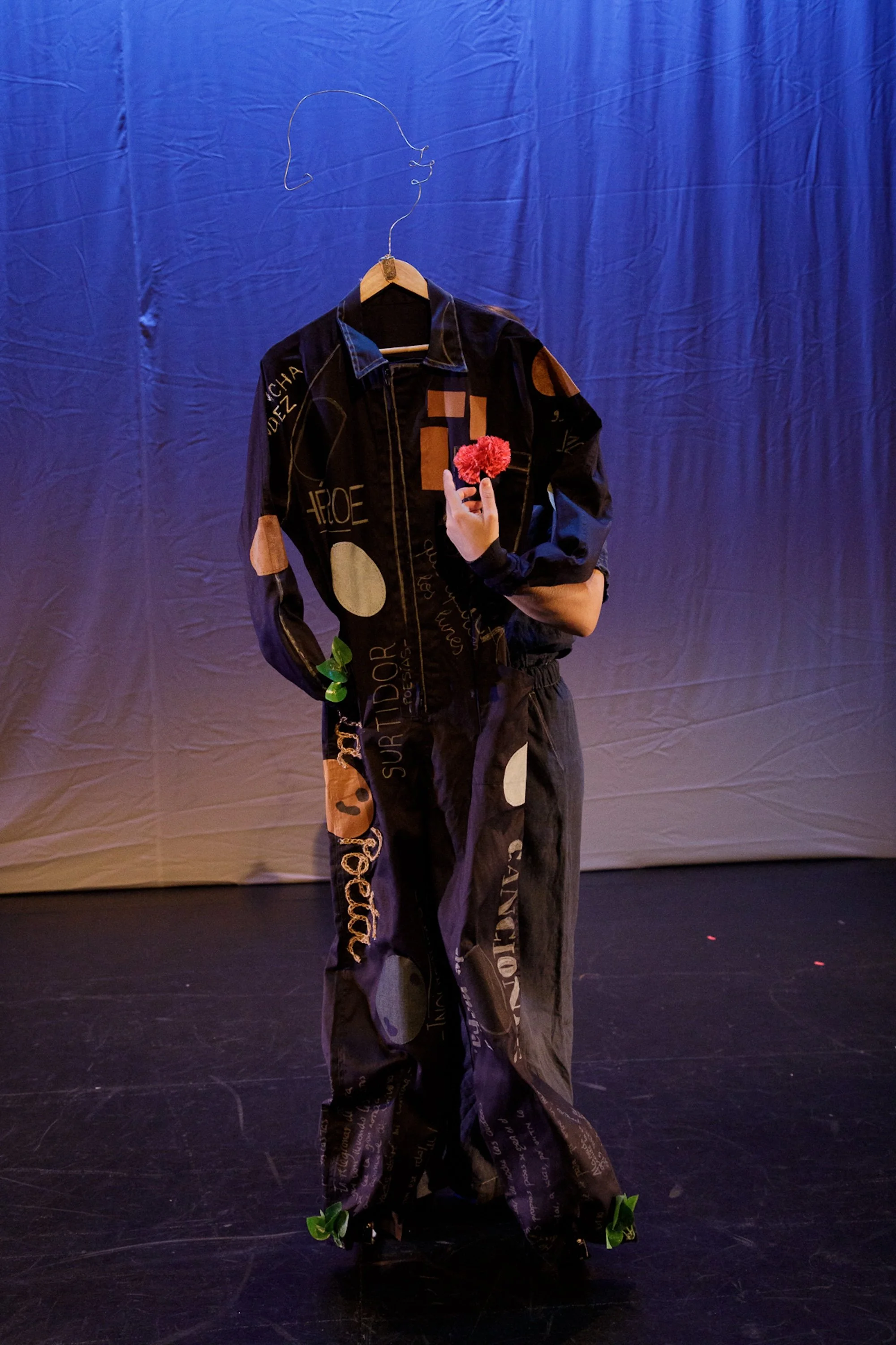 Ropa de moda con escritos y diseños, colgada en un perchero con una figura de alambre en forma de cabeza y manos con flores rojas, en un escenario con cortina azul.