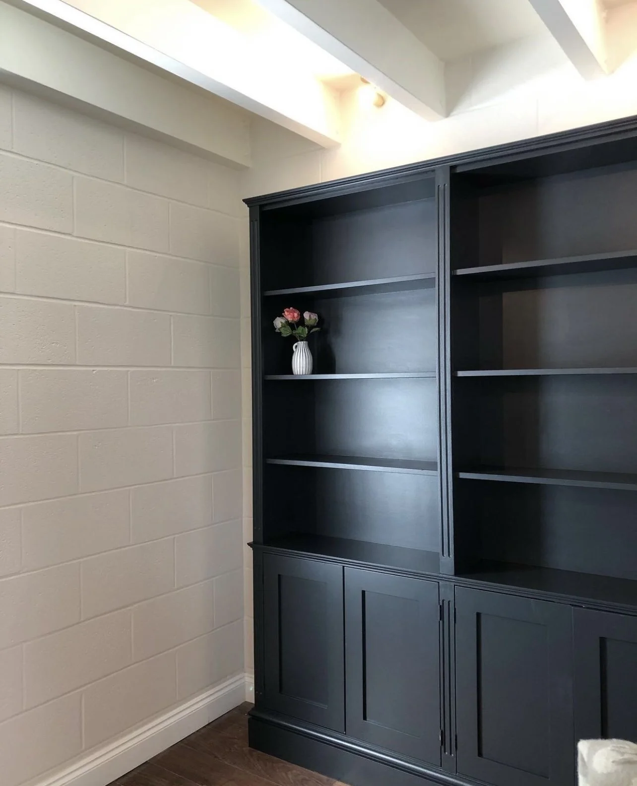 A black wooden bookshelf with several empty shelves and a small white vase with pink and red flowers on one of the shelves, against a beige brick wall with a corner of the ceiling visible.