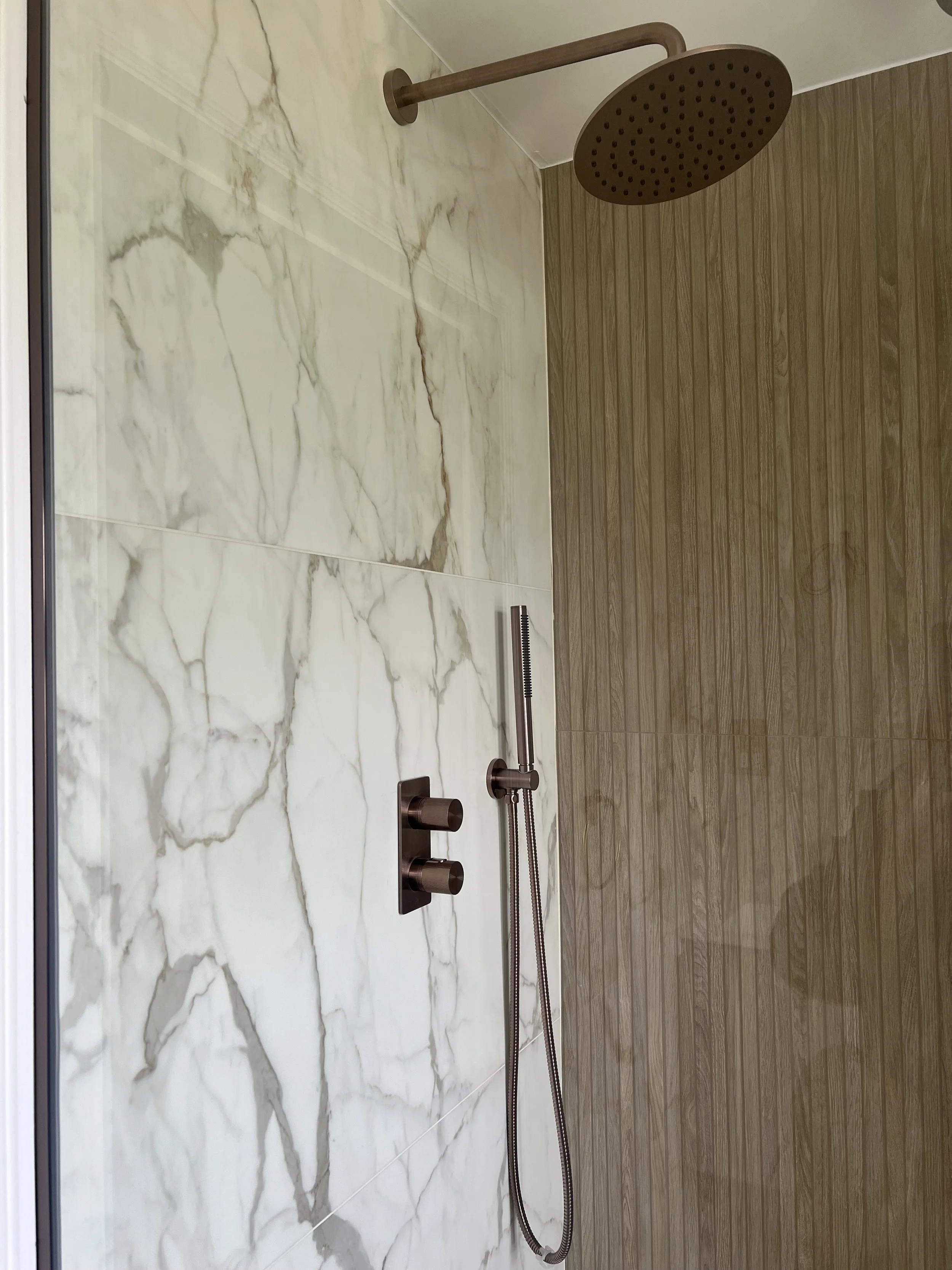 A modern shower area with a large round overhead shower head, a handheld shower with a hose, and controls on a wall that combines marble and wood-like tiles.
