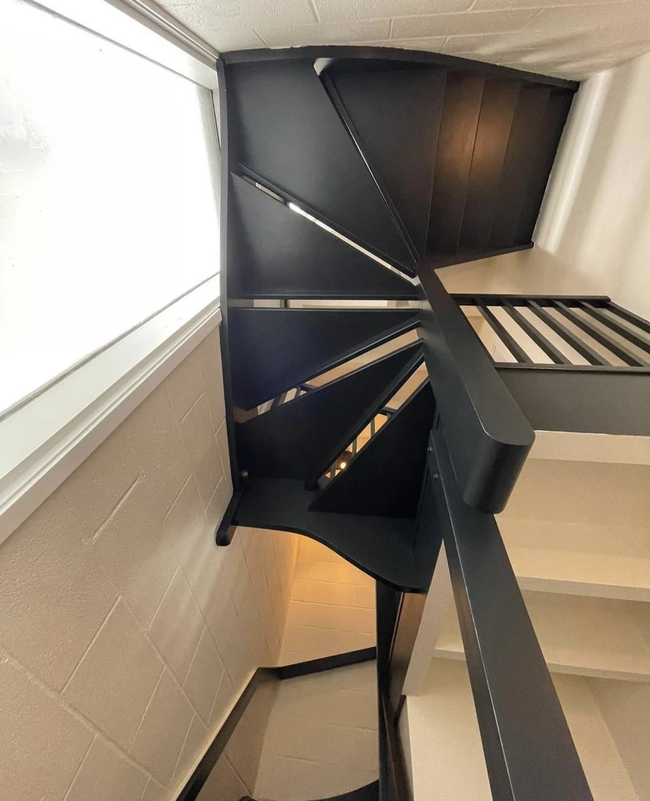 Black spiral staircase with wooden steps, viewed from the top near a window with white trim, inside a beige-walled room.