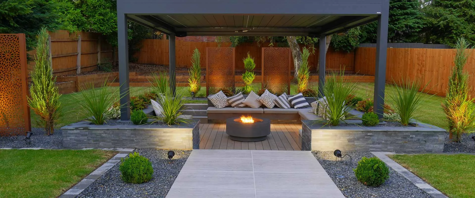 An outdoor patio area with a built-in curved seating area, decorative pillows, a fire pit in the center, and a pergola overhead. Surrounding the space are landscaped plants and a wooden fence.