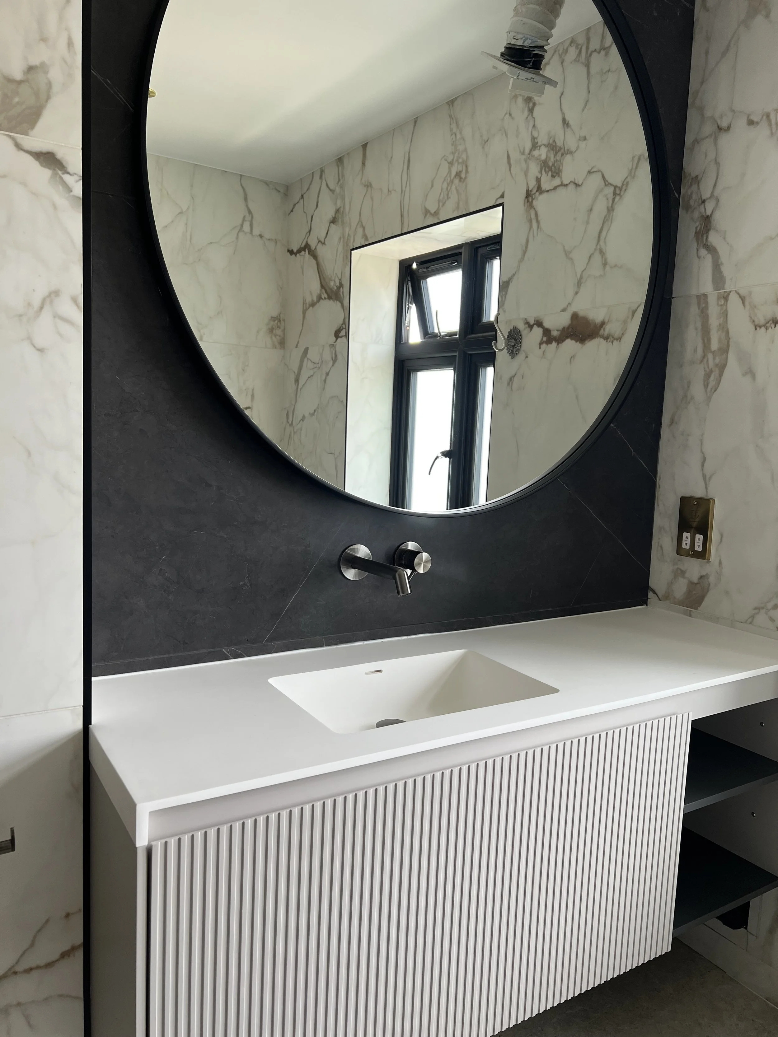 Modern bathroom vanity with a large round mirror, marble walls, a window, and a sink with a black wall behind it.