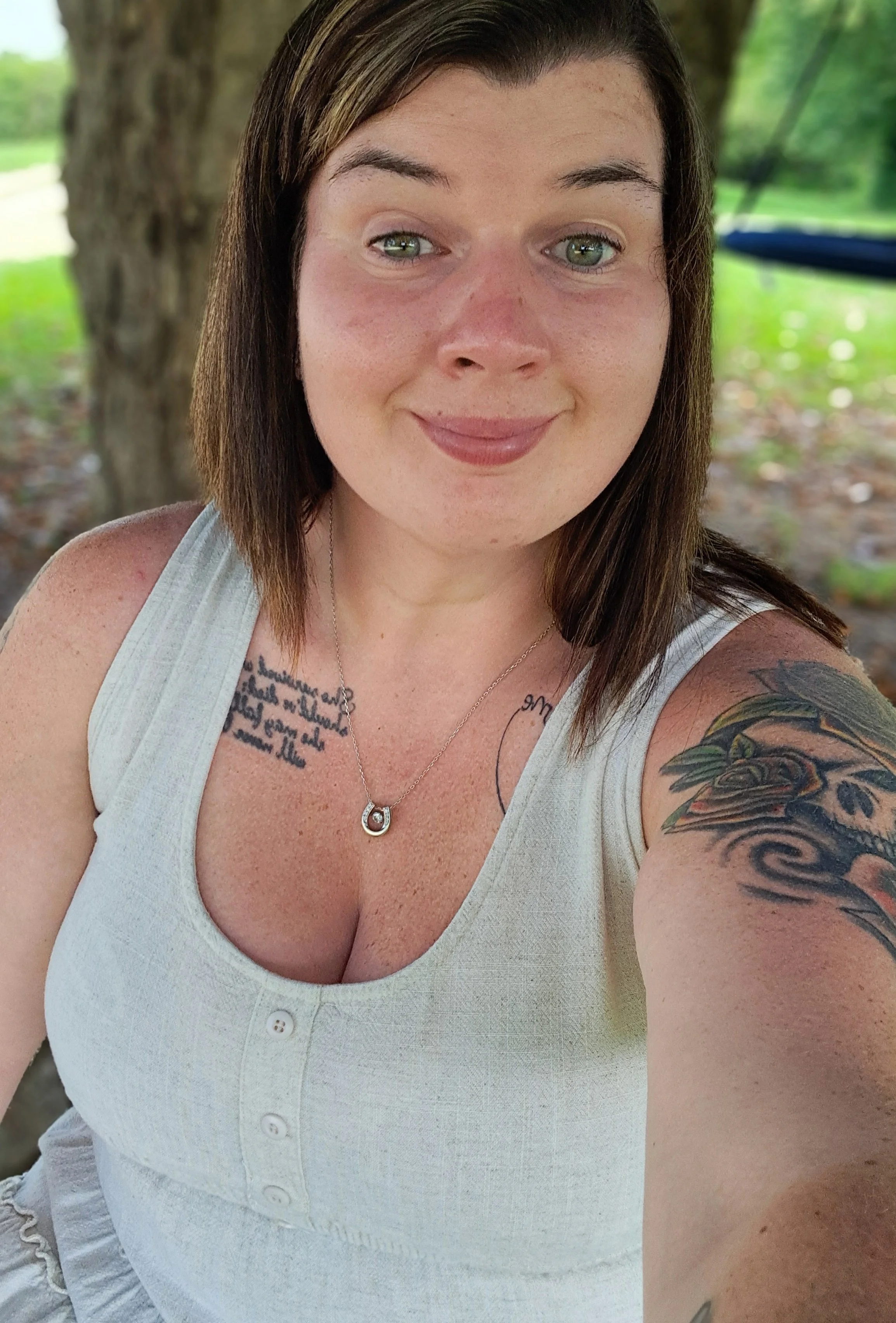 A smiling woman with shoulder-length brown hair, tattoos on her shoulder and chest, wearing a white sleeveless top and a necklace, sitting outdoors near a tree in a park.